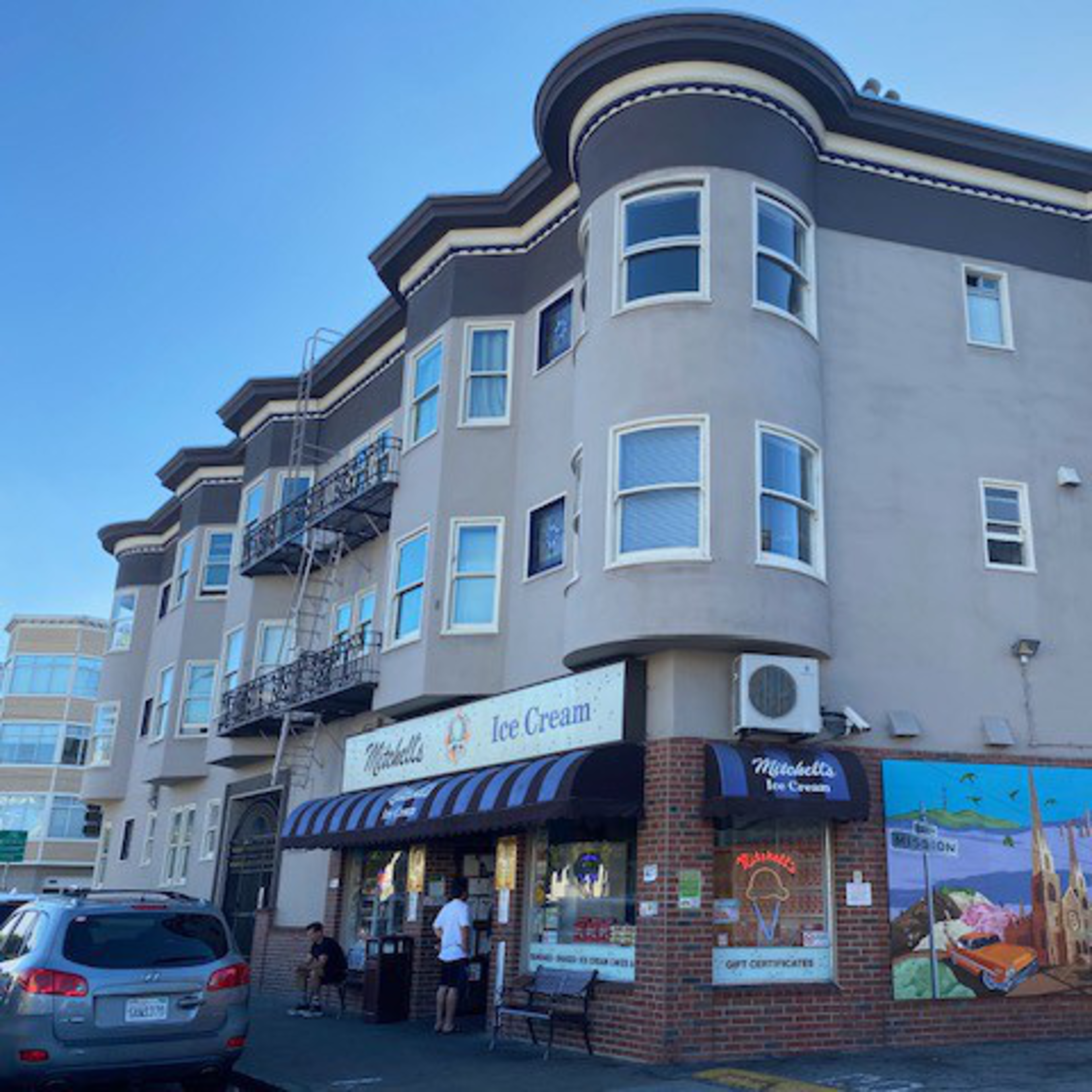 The image shows a beige, multi-story building with an ice cream shop on the ground floor. The shop has a sign and a striped awning, with a mural painted on the side wall.