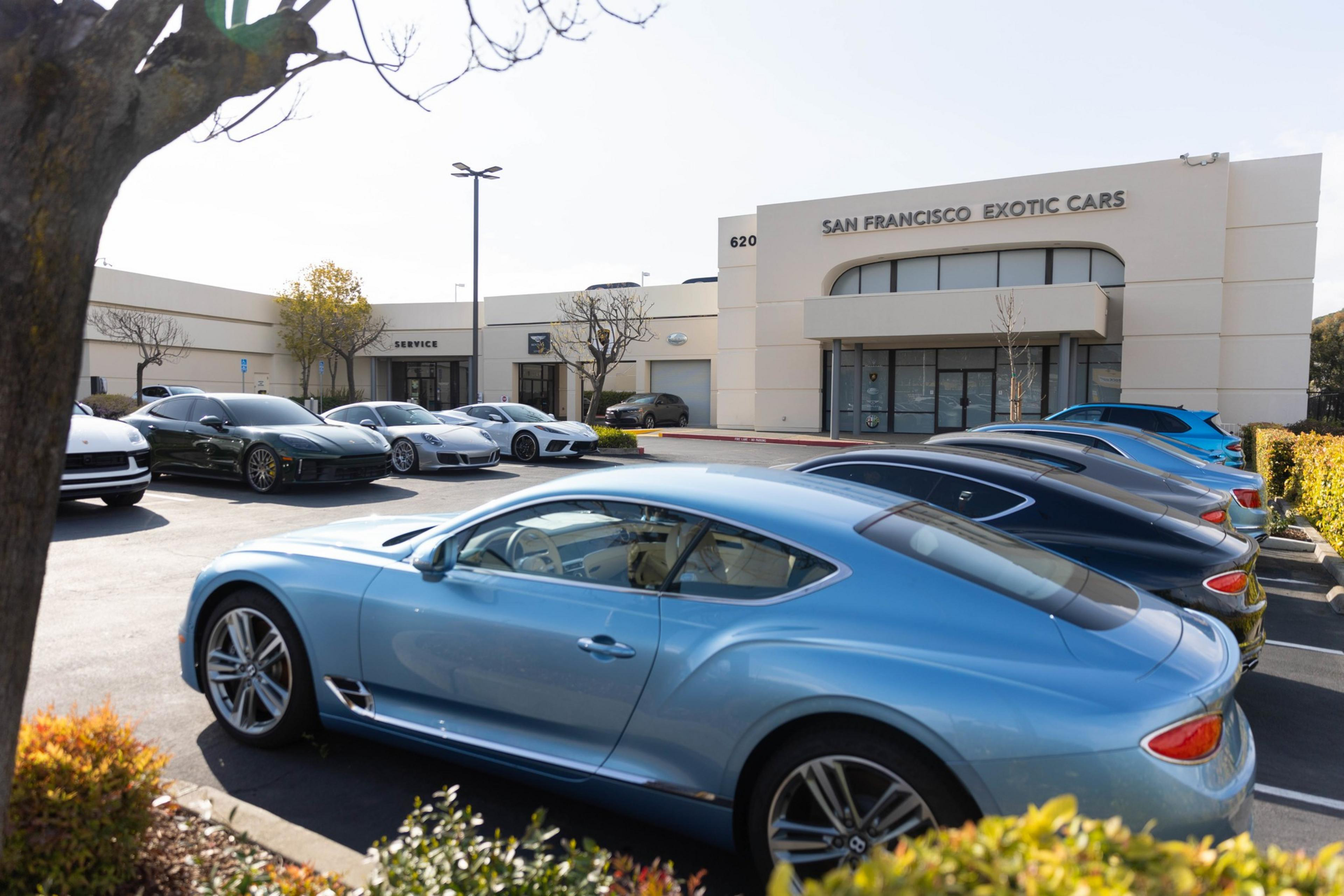 A row of luxury cars is parked outside a dealership named &quot;San Francisco Exotic Cars.&quot; The cars are sleek and polished in various colors.
