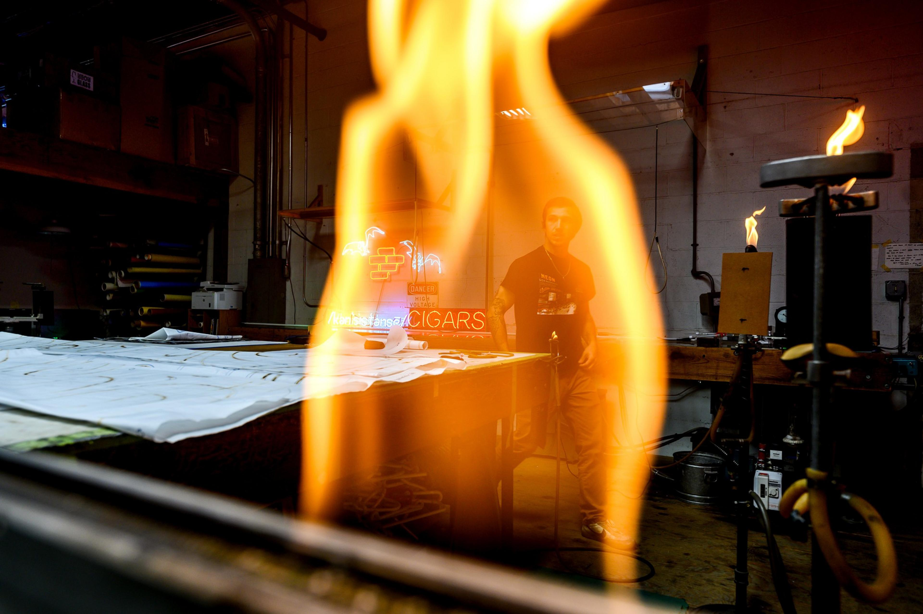 A large flame of fire in the foreground with a man in the background of a workshop.