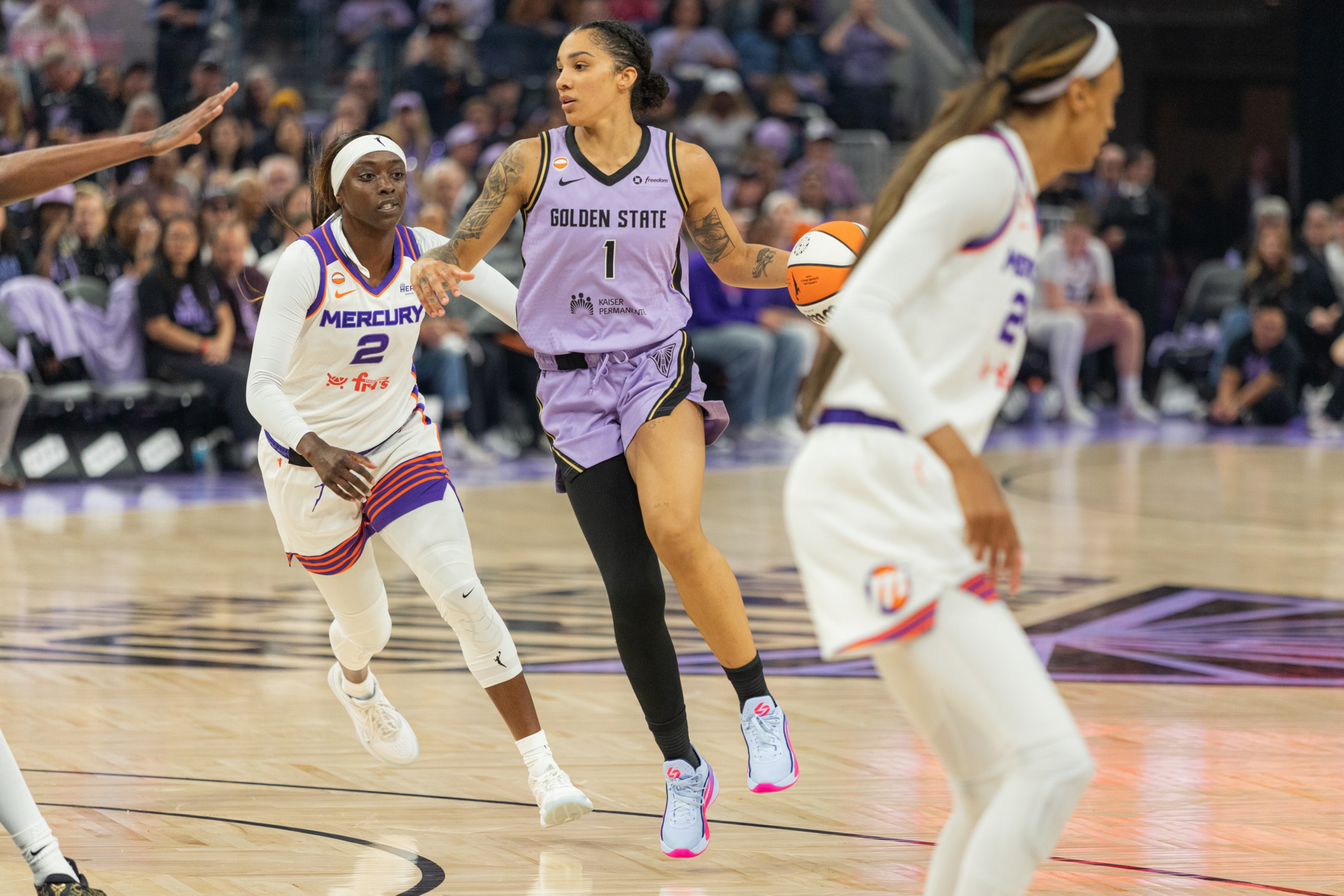 A basketball player in a purple Golden State uniform dribbles the ball while being closely guarded by players in white Phoenix Mercury uniforms.