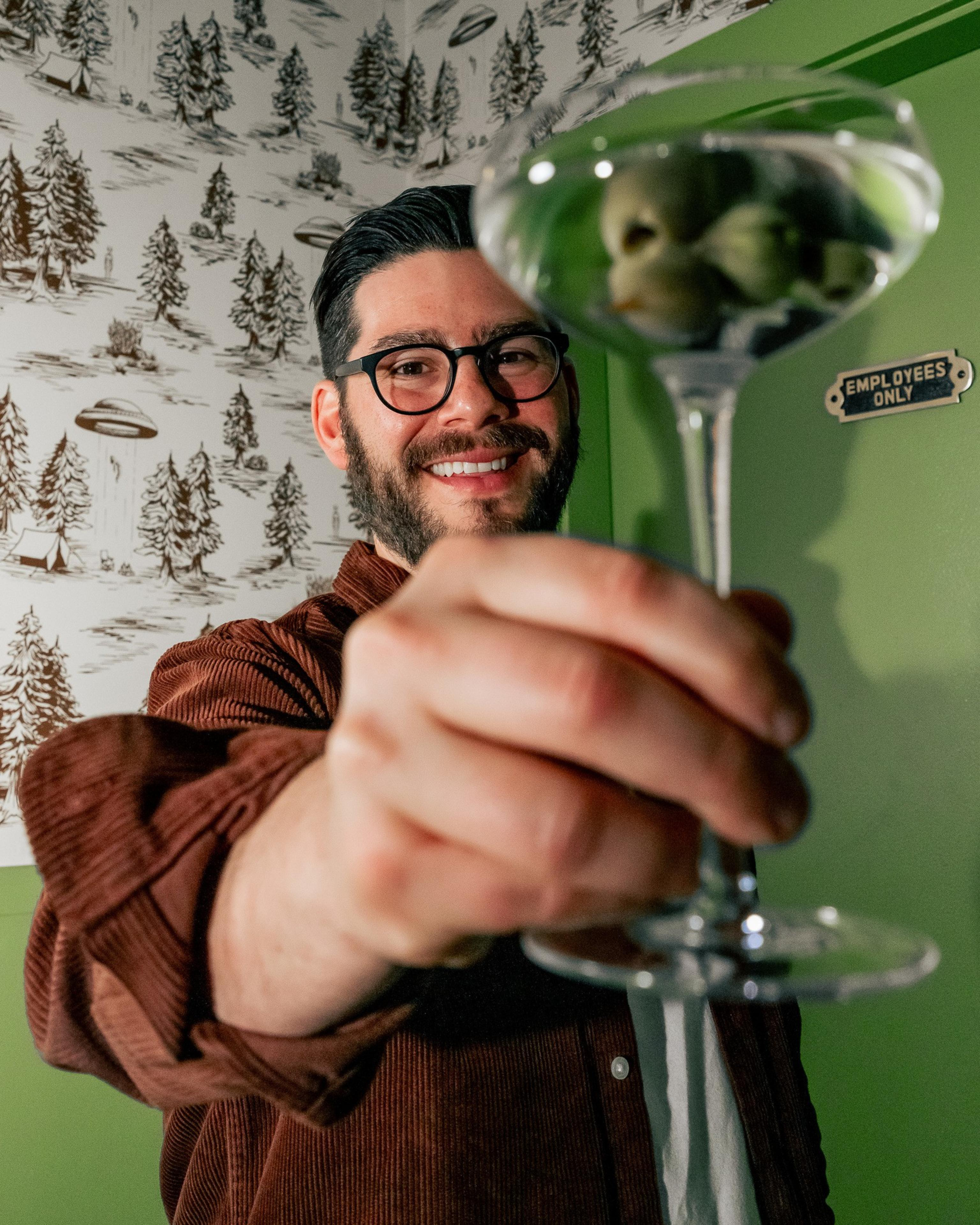 A person wearing glasses and a brown shirt holds a martini glass with olives. They're smiling, standing in front of a wallpaper with trees and UFOs.