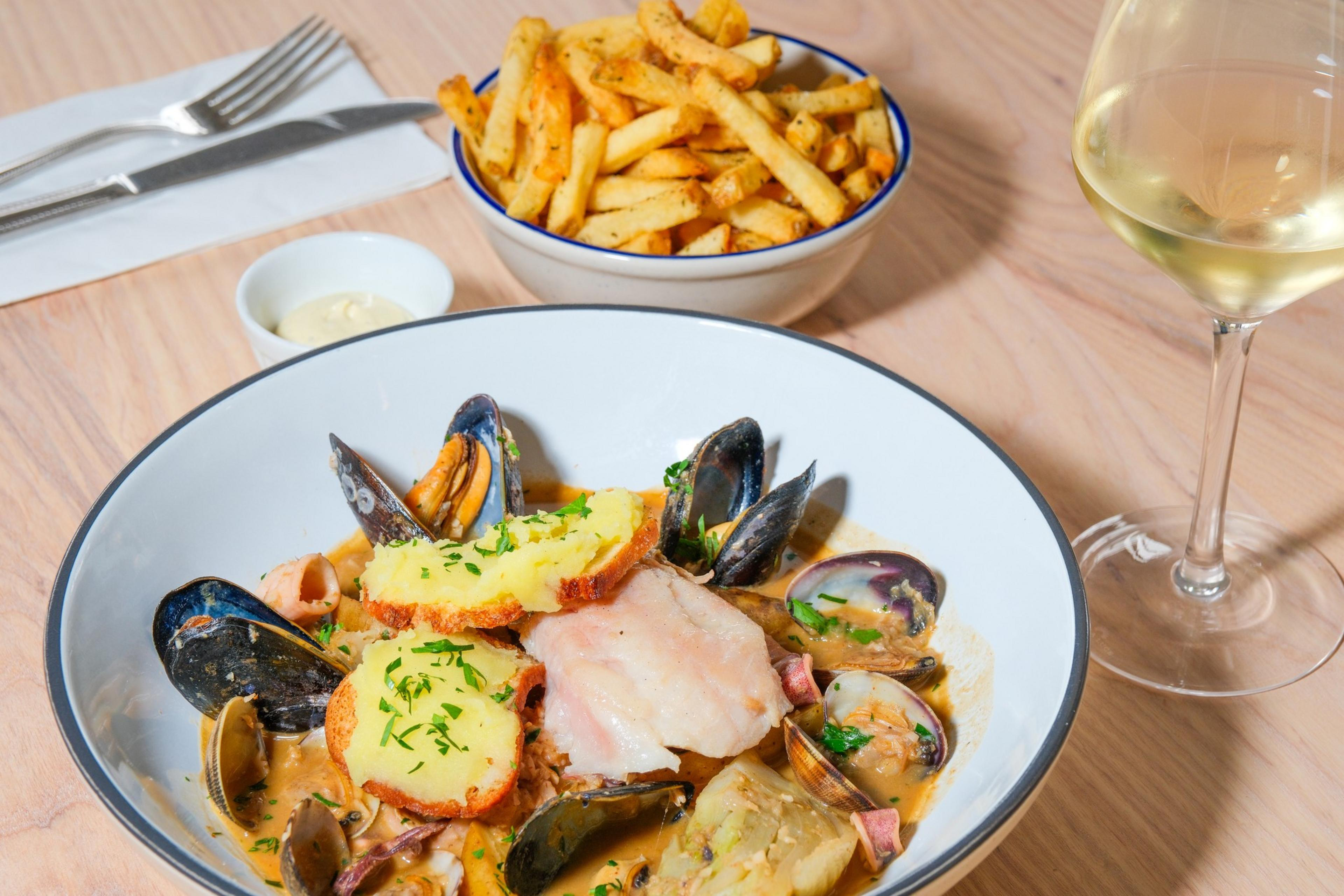 The image shows a bowl of seafood stew with mussels, clams, and fish, accompanied by seasoned fries, a small dish of sauce, and a glass of white wine on a wooden table.