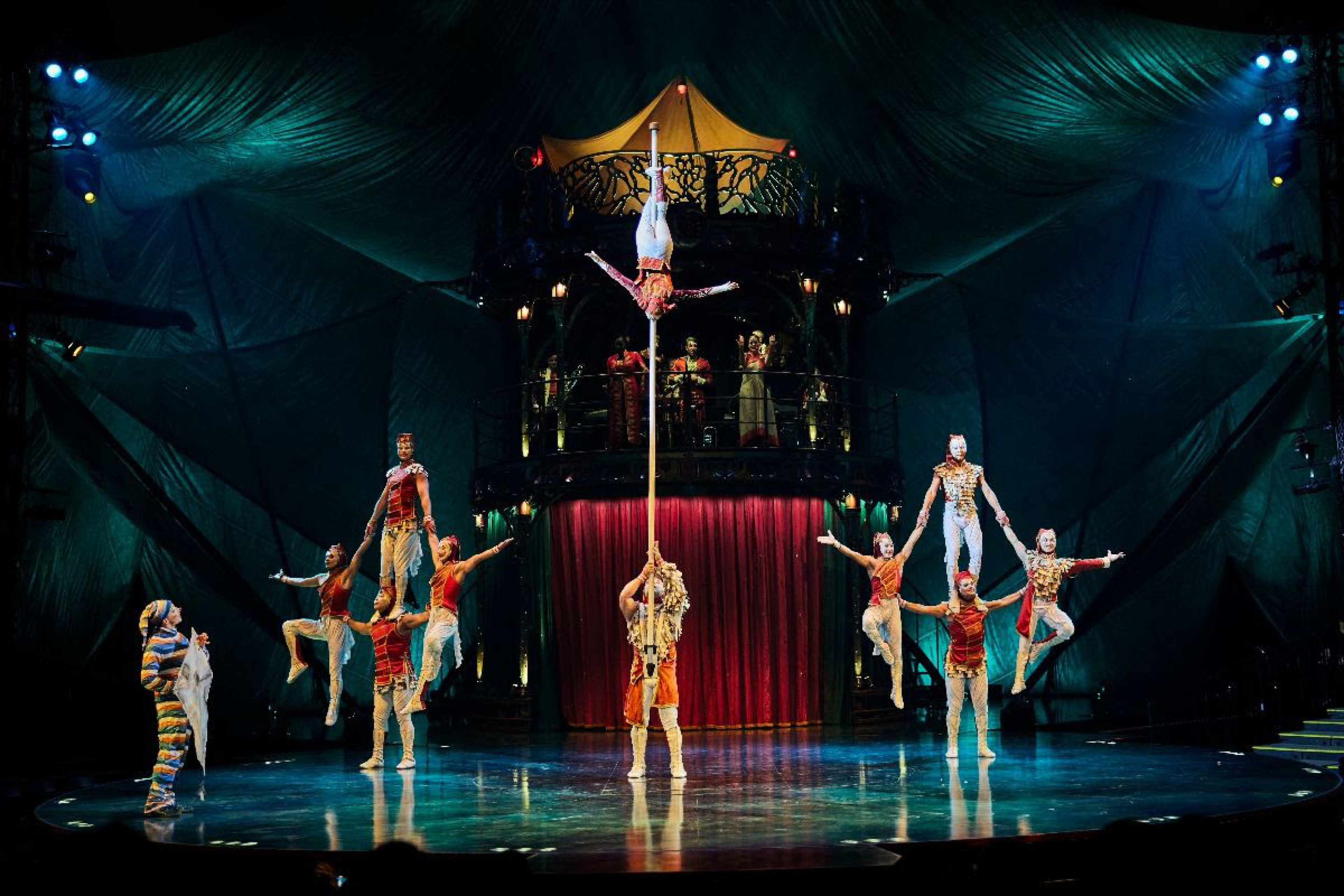 Performers in colorful costumes balance in acrobatic poses on a stage with dramatic lighting and a tall pole supporting an inverted aerialist.
