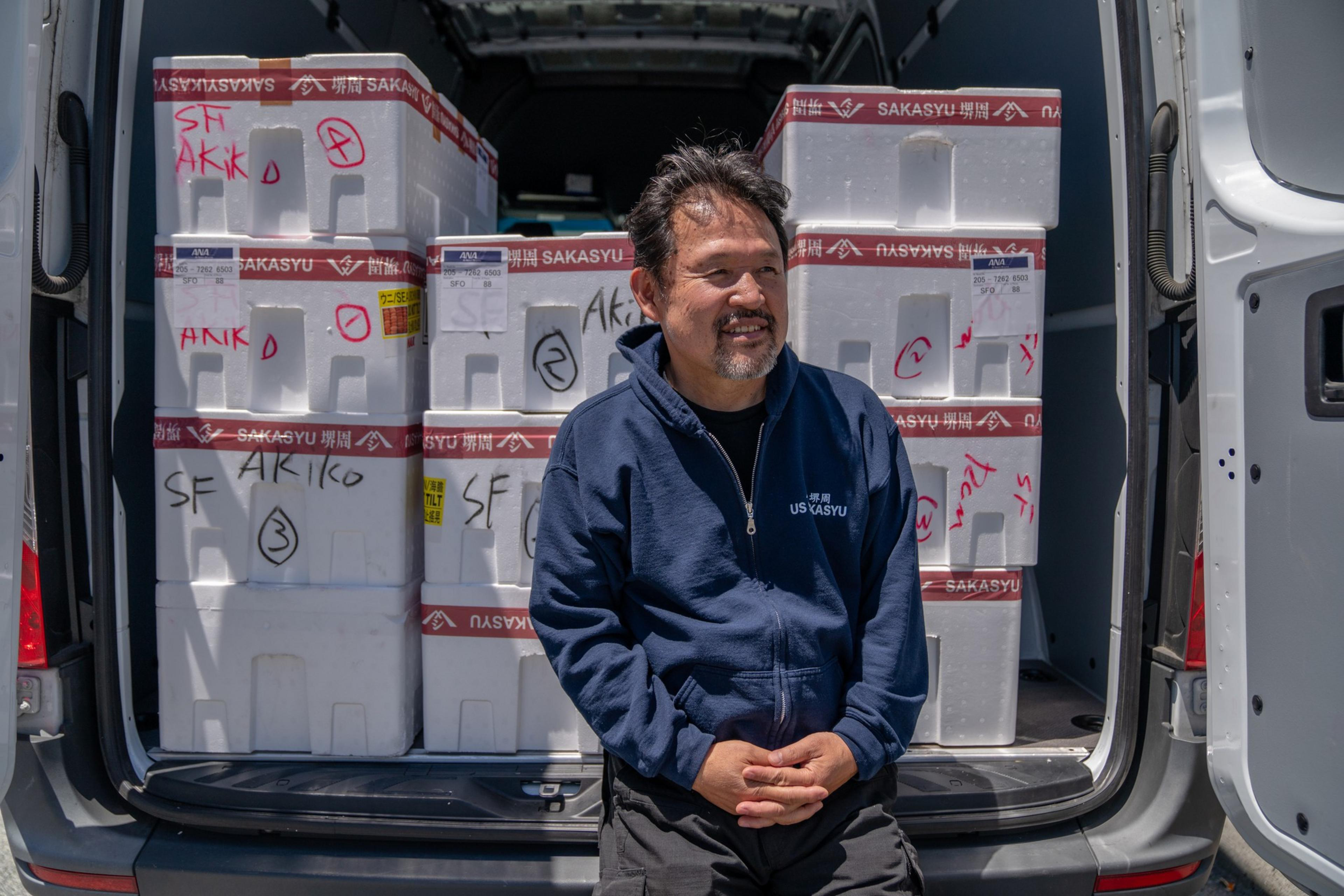 A man in a blue hoodie stands beside an open van filled with labeled white containers stacked neatly; he is smiling and looking to the side.