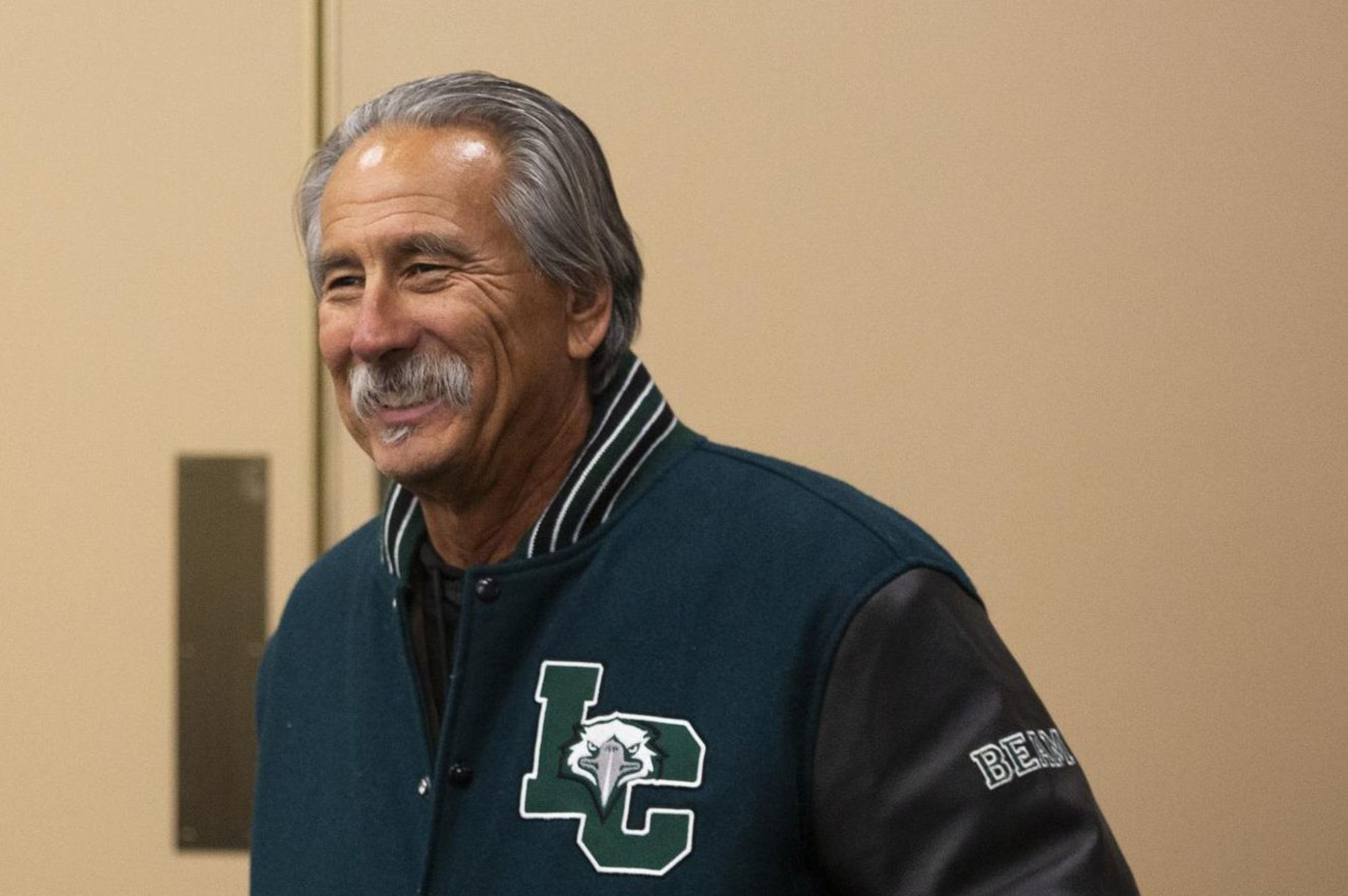 An older man with gray hair and a mustache smiles while wearing a dark green and black letterman jacket with "LC" and an eagle logo on it.