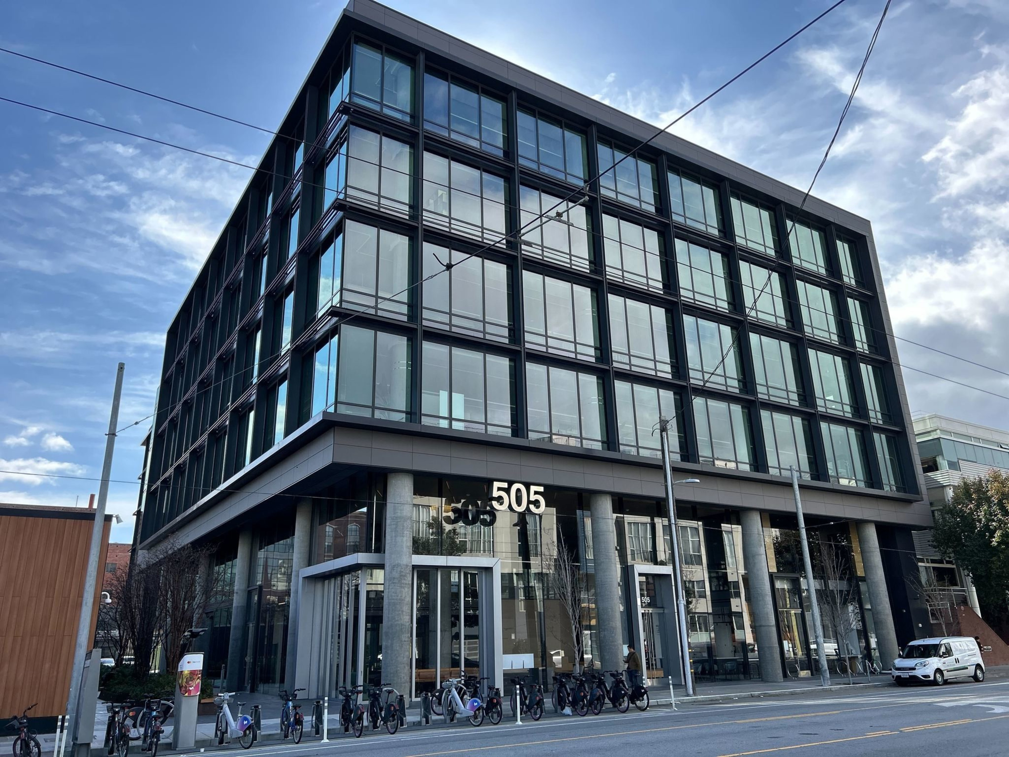 Modern building with large windows under a blue sky, number 505 on the facade, and parked bicycles in front.
