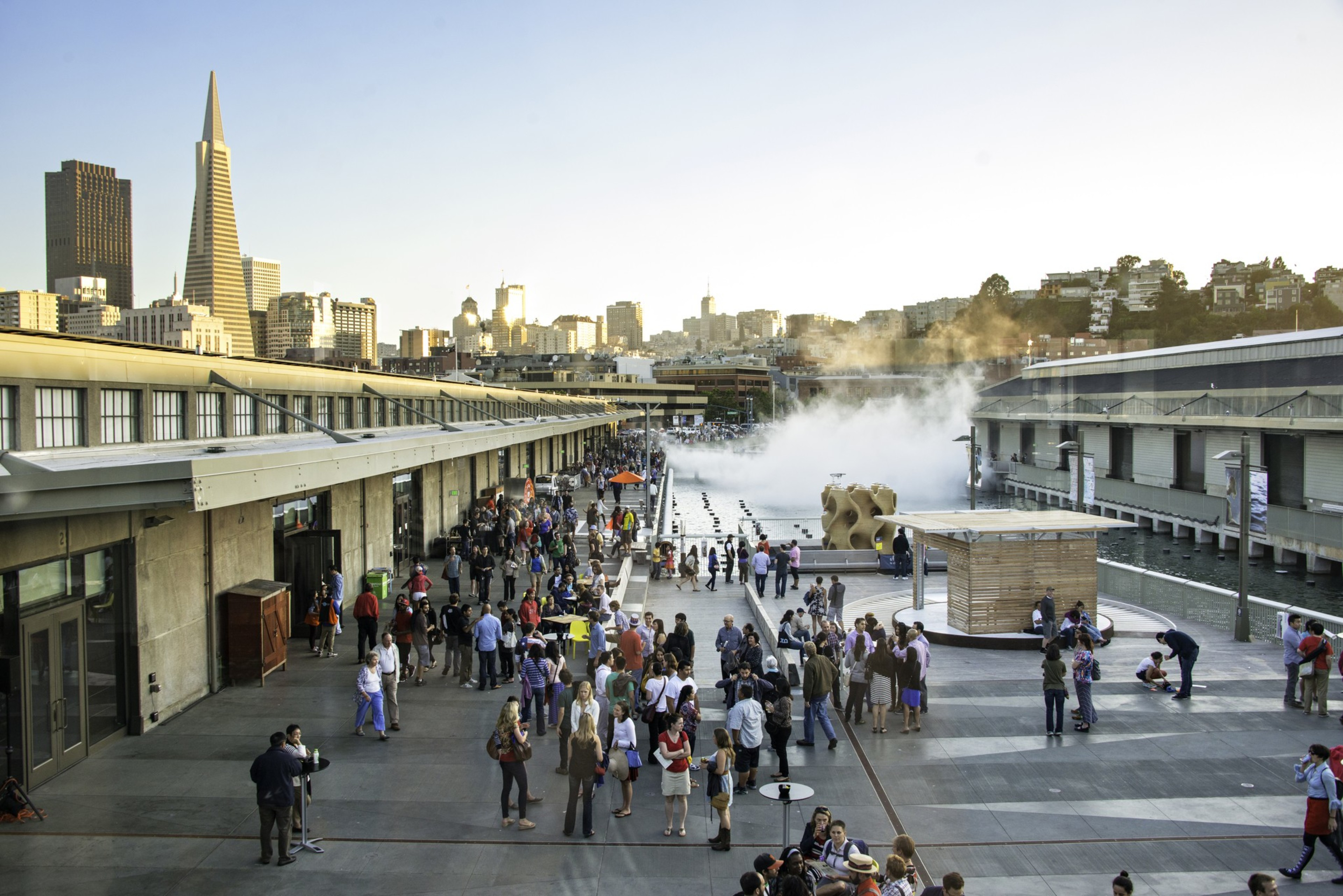 A bustling outdoor waterfront scene with many people gathered on a wide promenade. Skyscrapers are visible in the background, and mist rises near the docks.