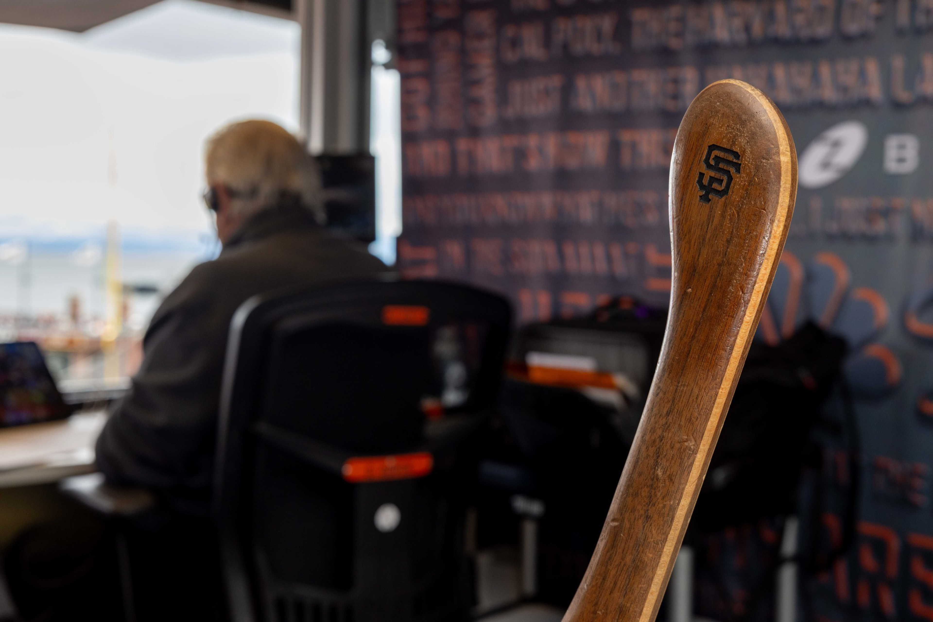 A wooden baseball bat with the San Francisco Giants logo leans in the foreground, while a person with headphones sits at a desk in the background.