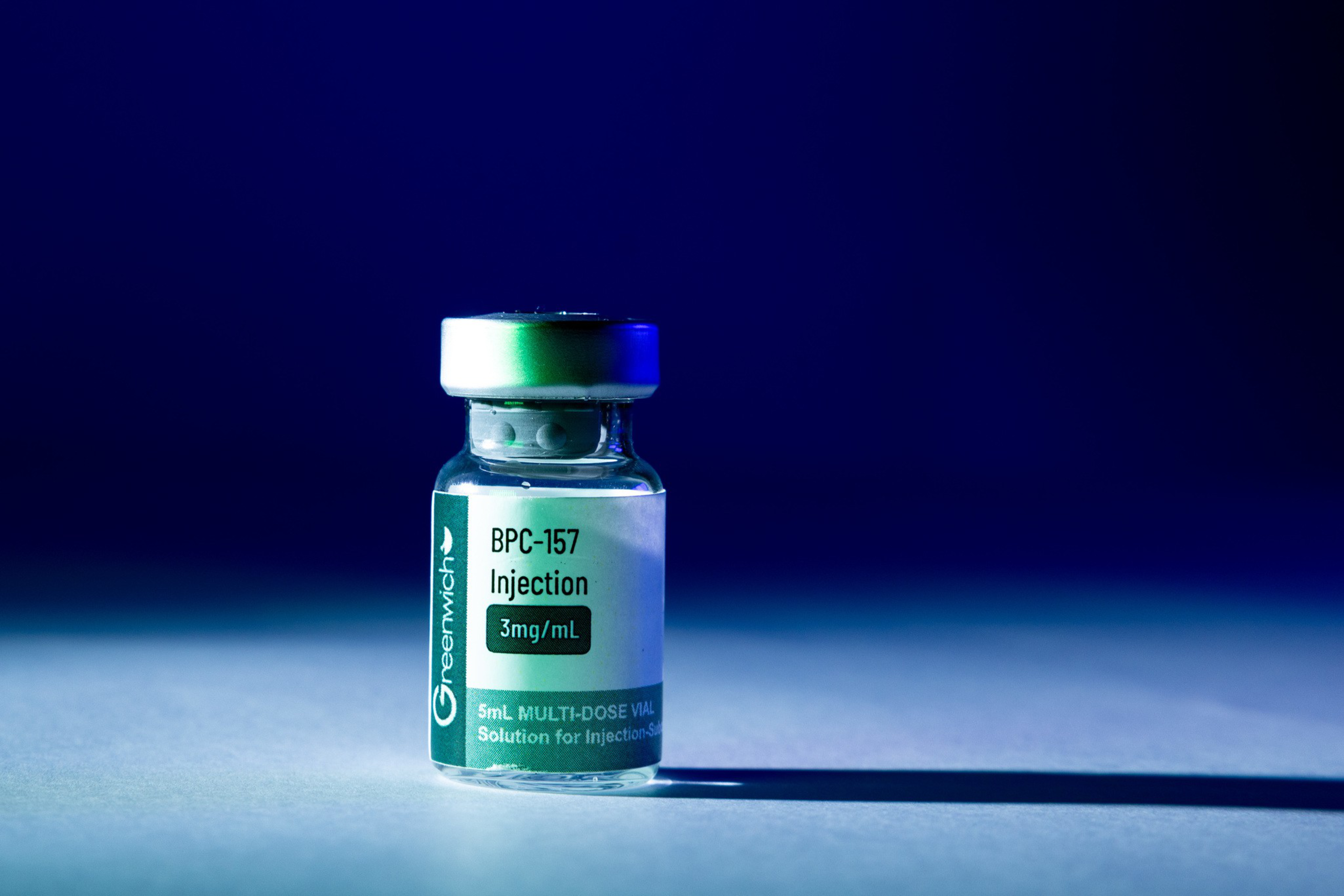 A small clear vial labeled “BPC-157 Injection 3mg/mL” from Greenwich, containing 5mL multi-dose solution for injection, is illuminated against a dark blue background.