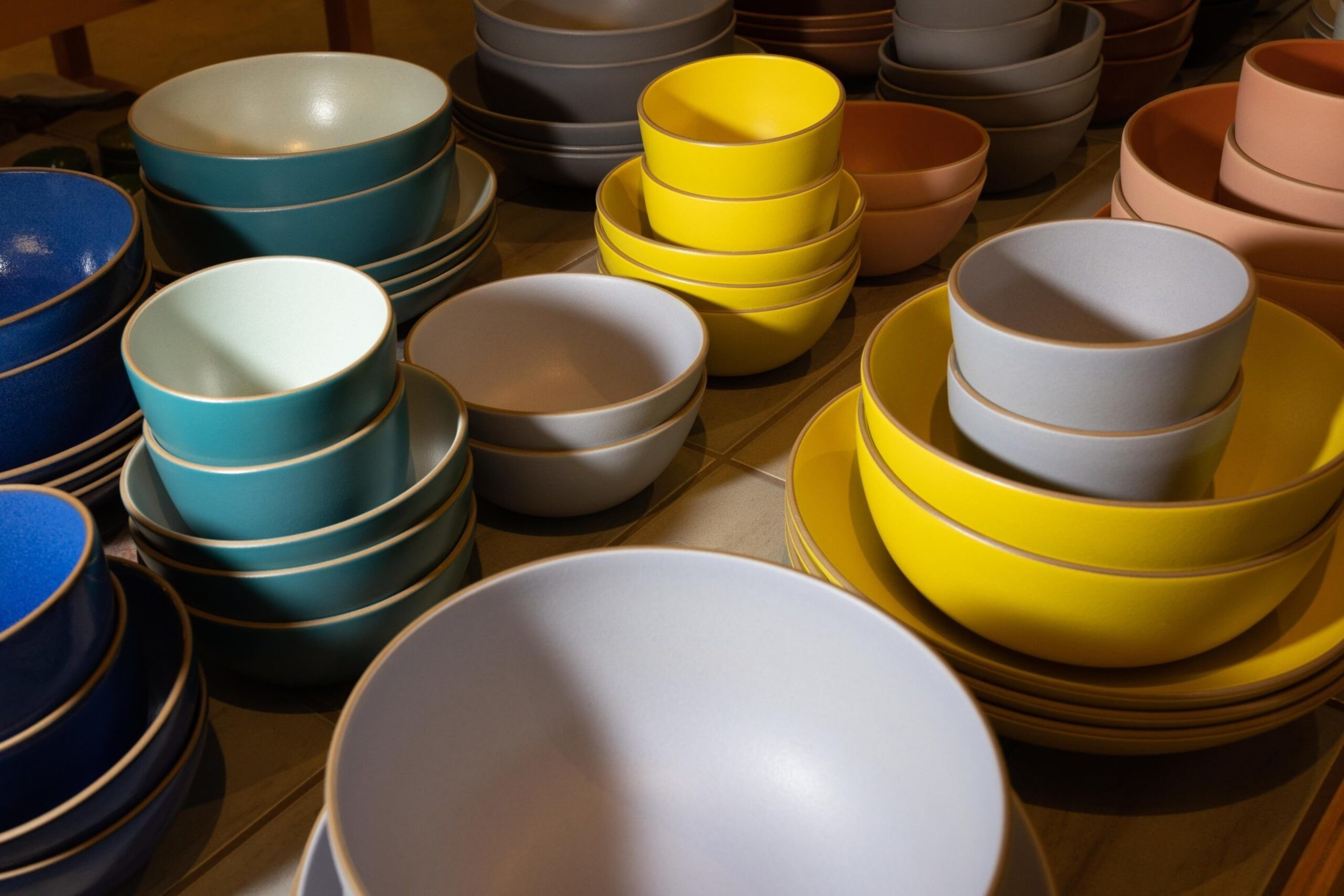 Stacks of ceramic bowls and plates in blue, teal, gray, yellow, and brown are arranged on a surface, illuminated by warm light.