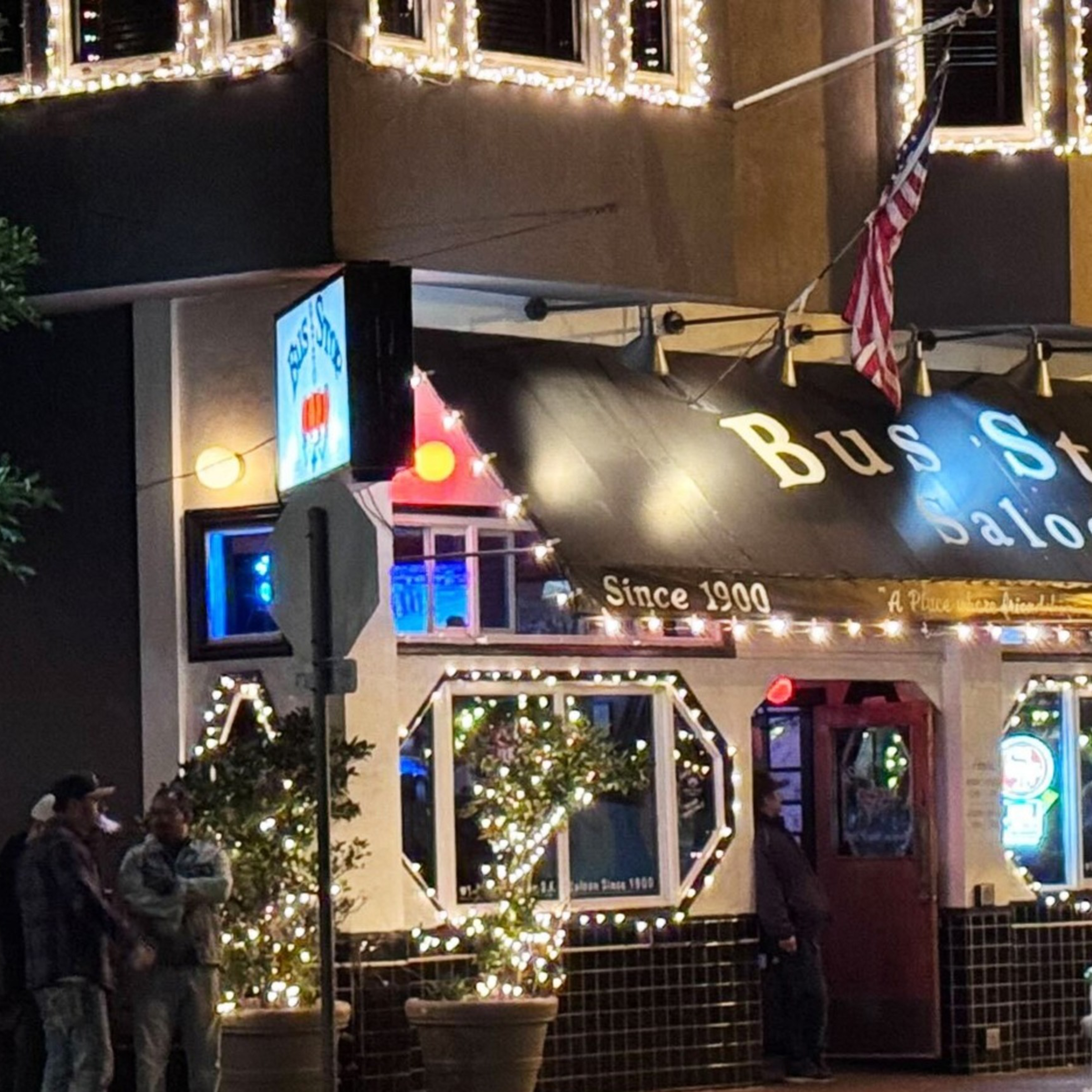 The image shows a warmly lit saloon named "Bus Stop" decorated with string lights. People are walking by on a crosswalk in front of the building at night.