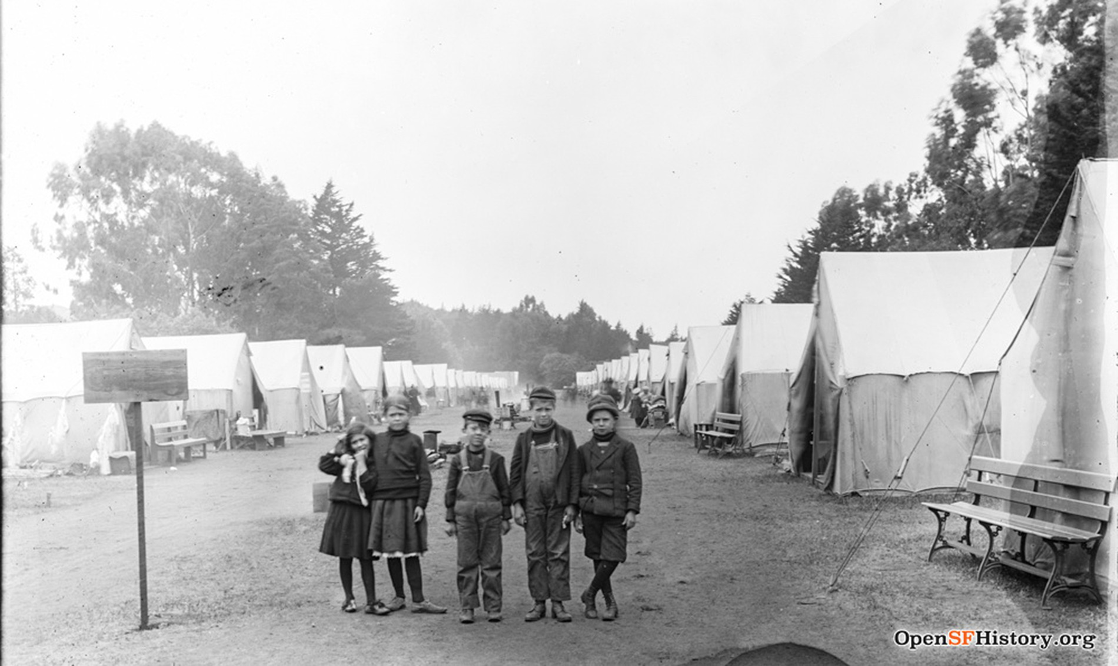 Five children stand on a dirt path between two rows of canvas tents in a camp surrounded by trees, with benches and a signpost visible.