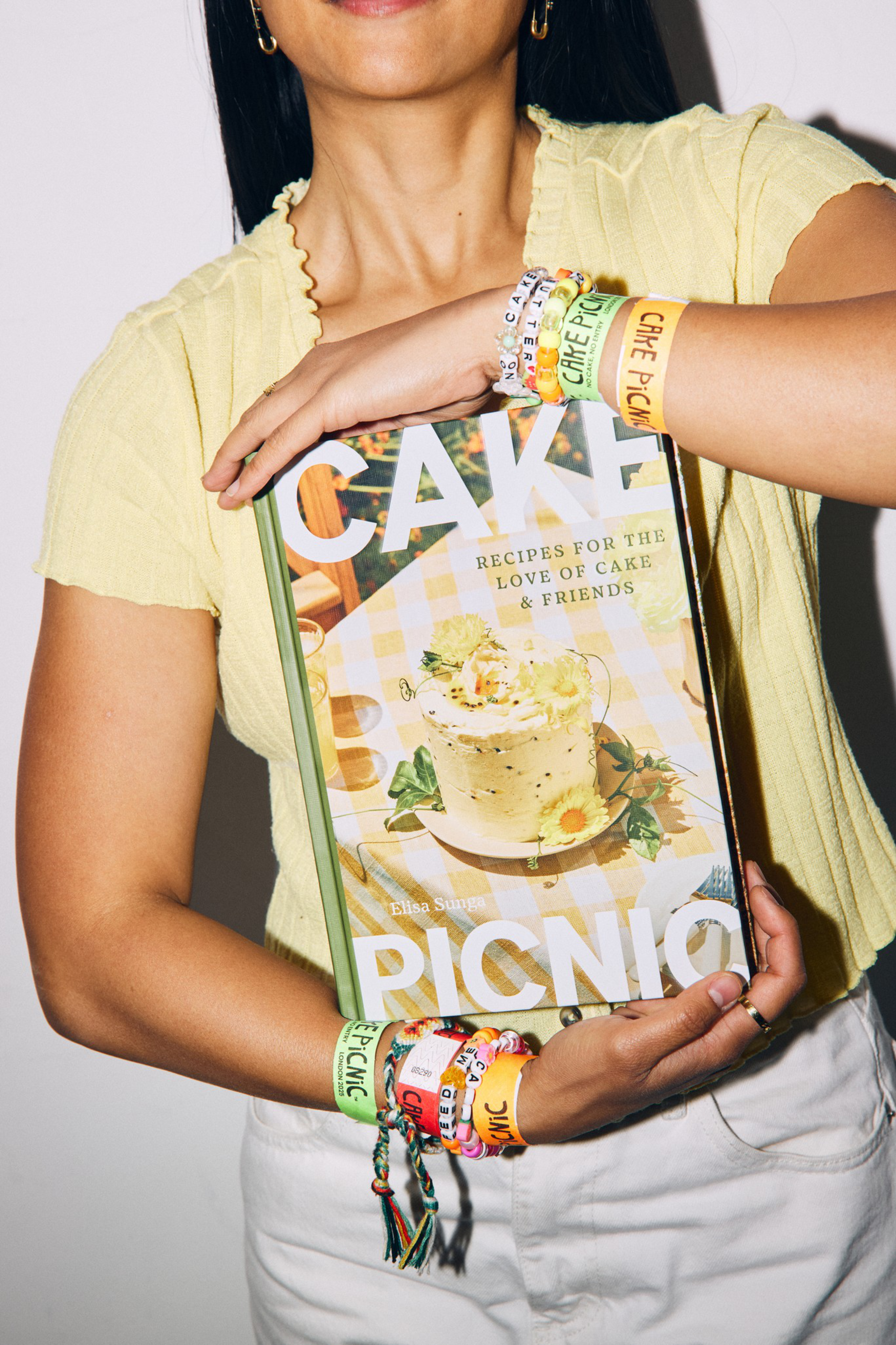 A person wearing a yellow top holds a cookbook titled “Cake Picnic,” adorned with multiple colorful bracelets that say “Cake Picnic.”