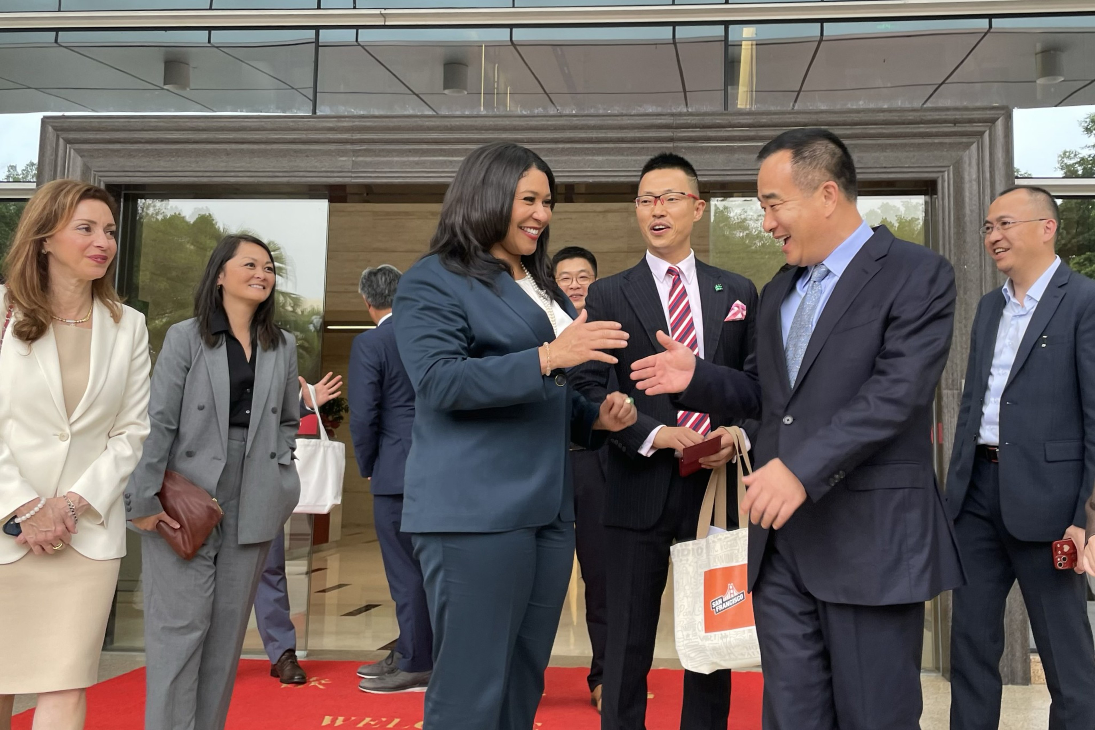 A group of smiling people exchanging gifts on a red carpet outside a modern building.