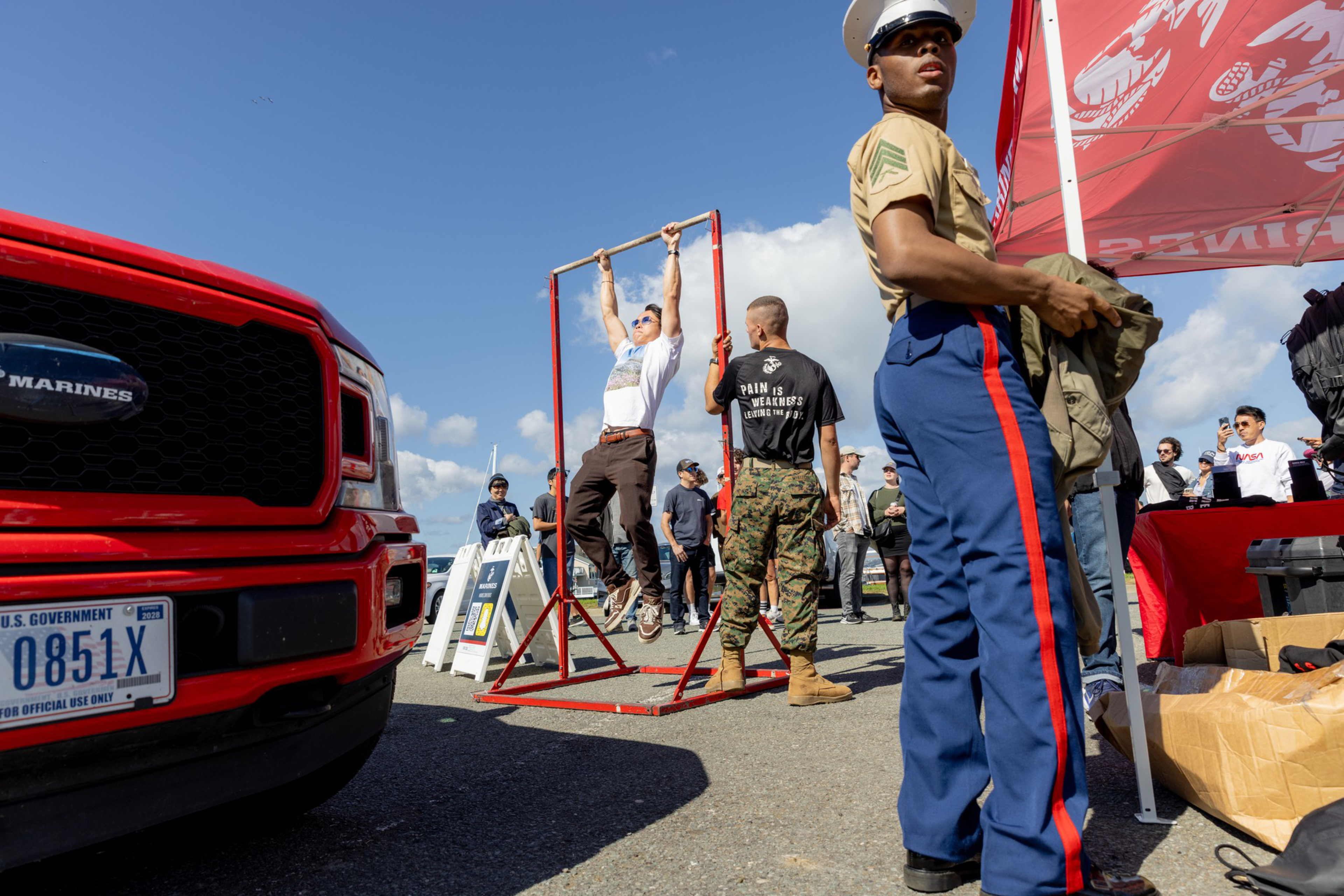 A man does pull-ups on a red bar while a Marine in uniform looks on near a red truck with a Marines logo, with people gathered around outside on a sunny day.