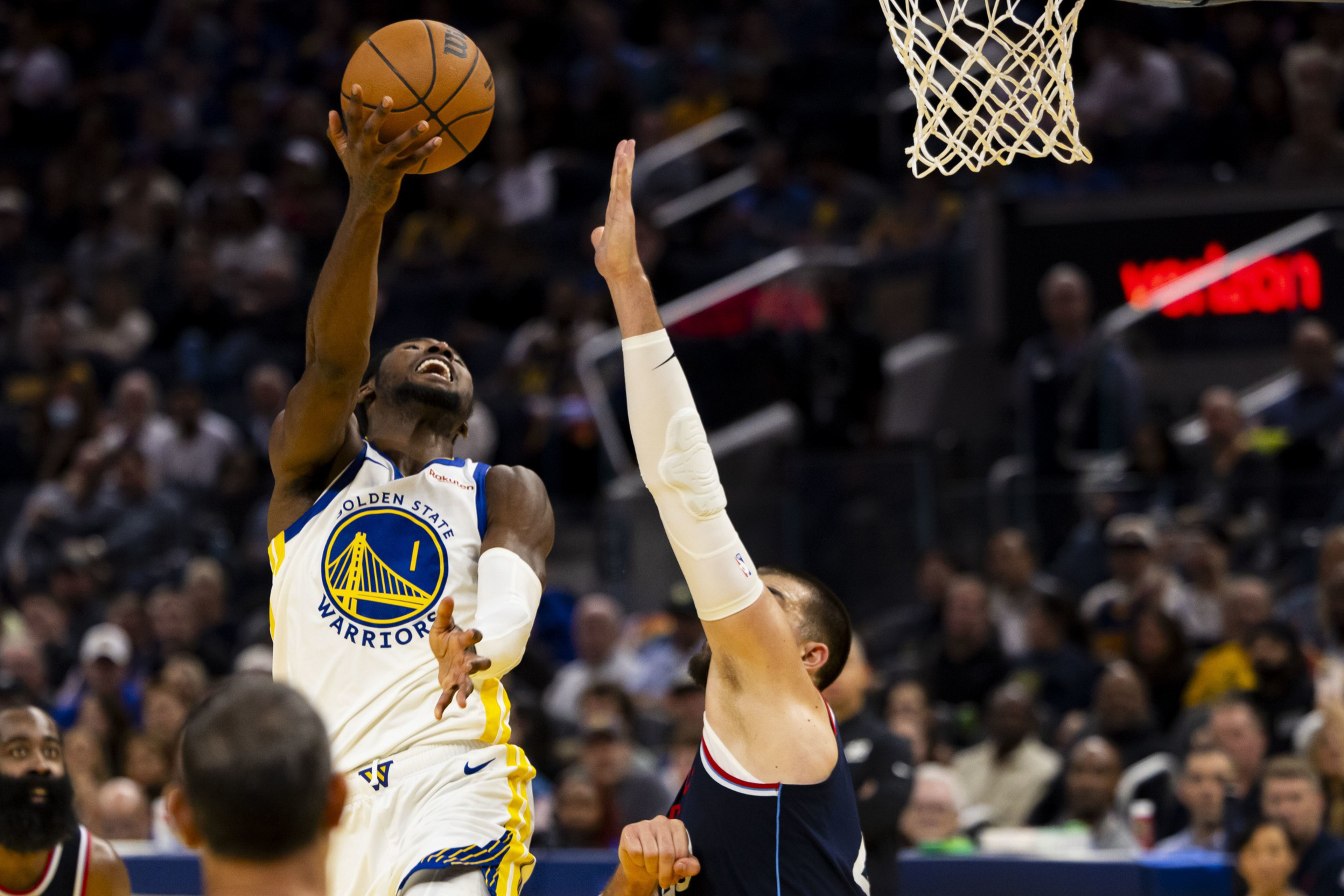 A Golden State Warriors player leaps for a layup as a defender with an outstretched arm tries to block the shot near the basketball hoop.