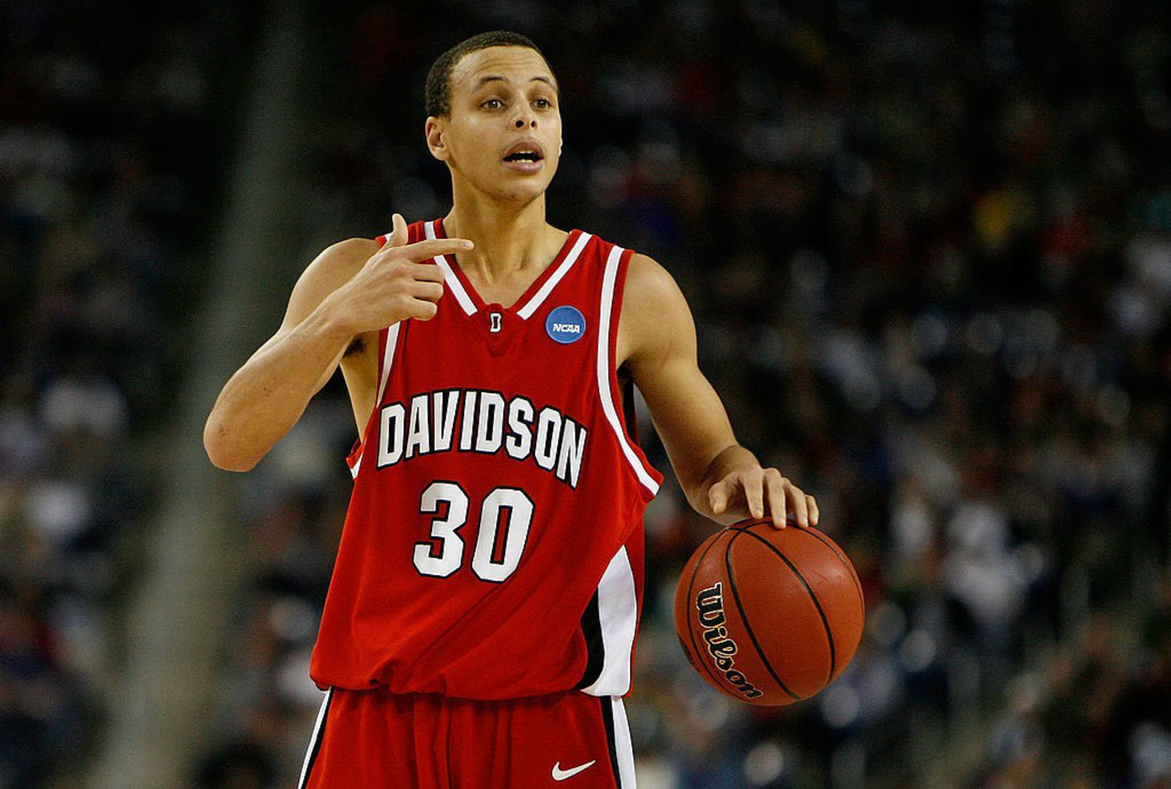 A basketball player in a red Davidson jersey with the number 30 holds the ball, gesturing with his left hand. The background shows a blurred audience.