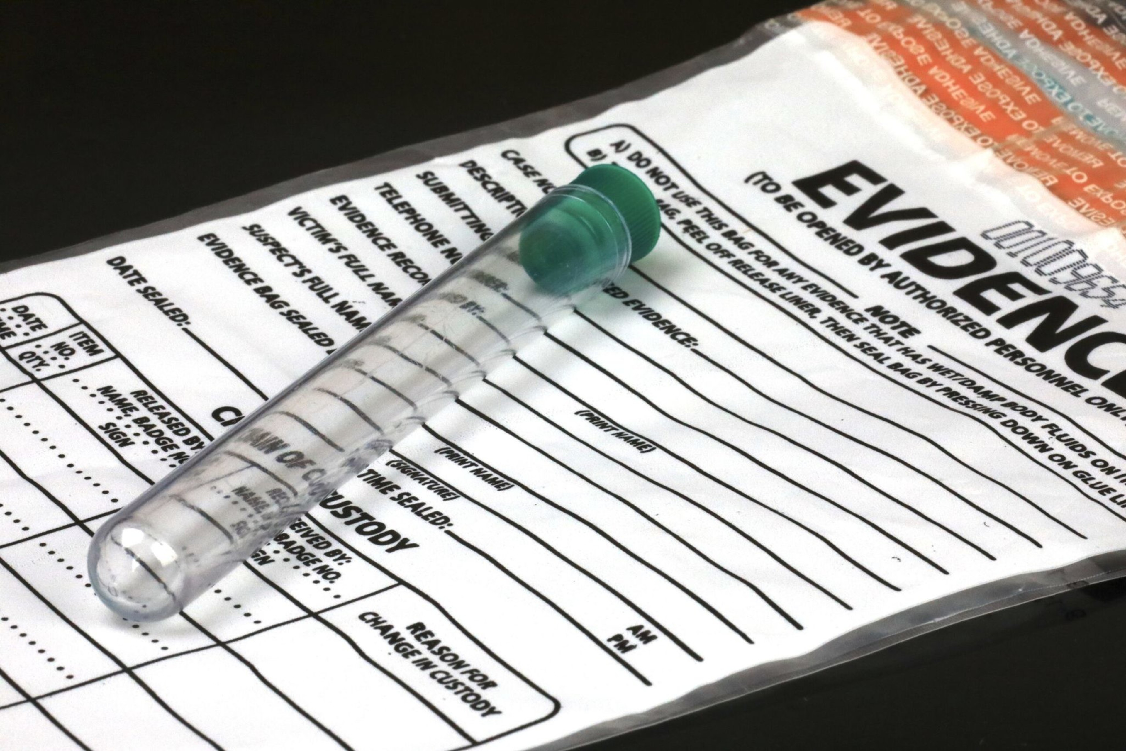 A sealed evidence bag lies flat with a clear plastic test tube capped in green placed on top, both on a dark surface.