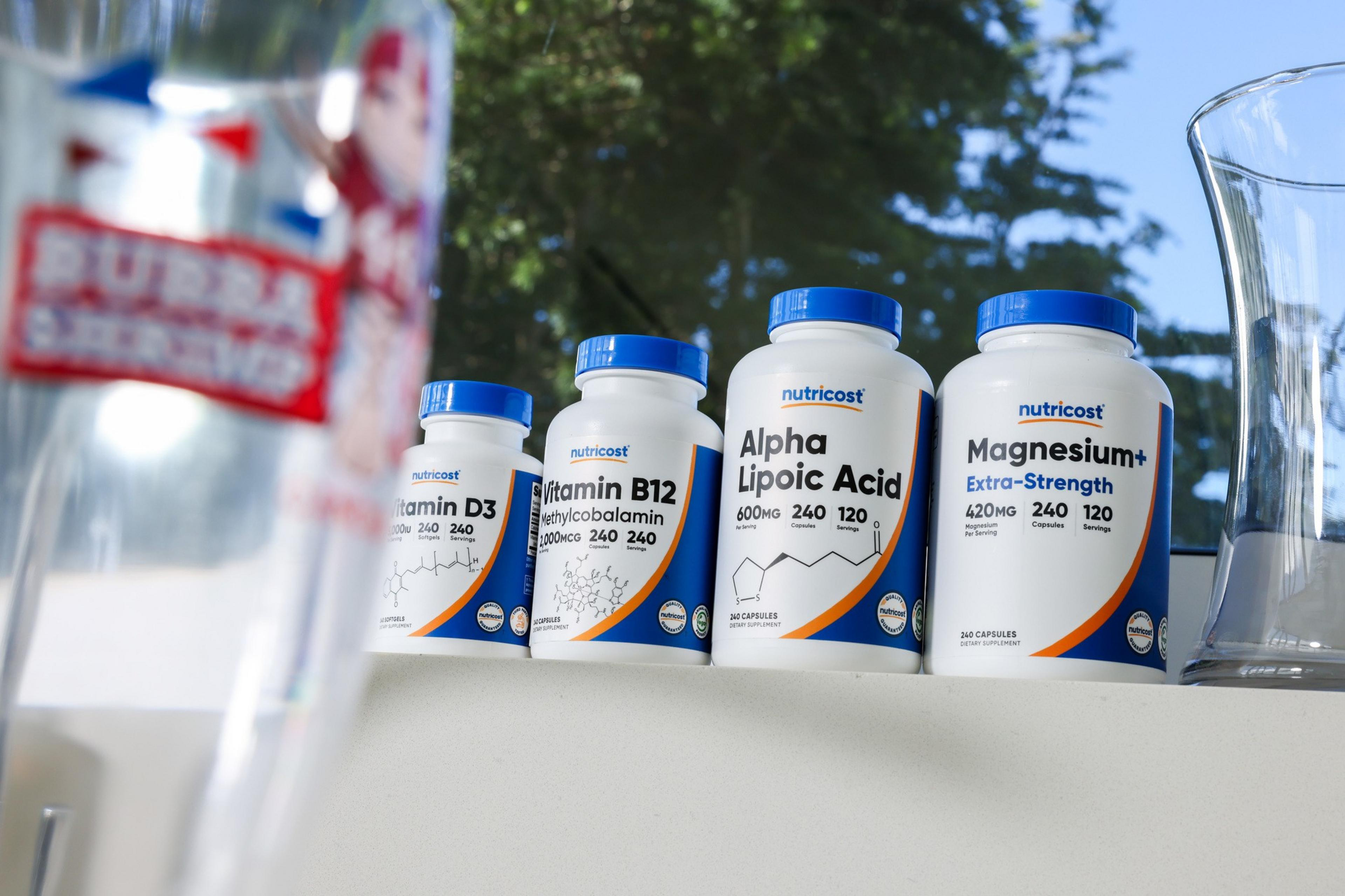 Four Nutricost supplement bottles with blue caps are lined up: Vitamin D3, Vitamin B12, Alpha Lipoic Acid, and Magnesium+, against a blurry tree background.
