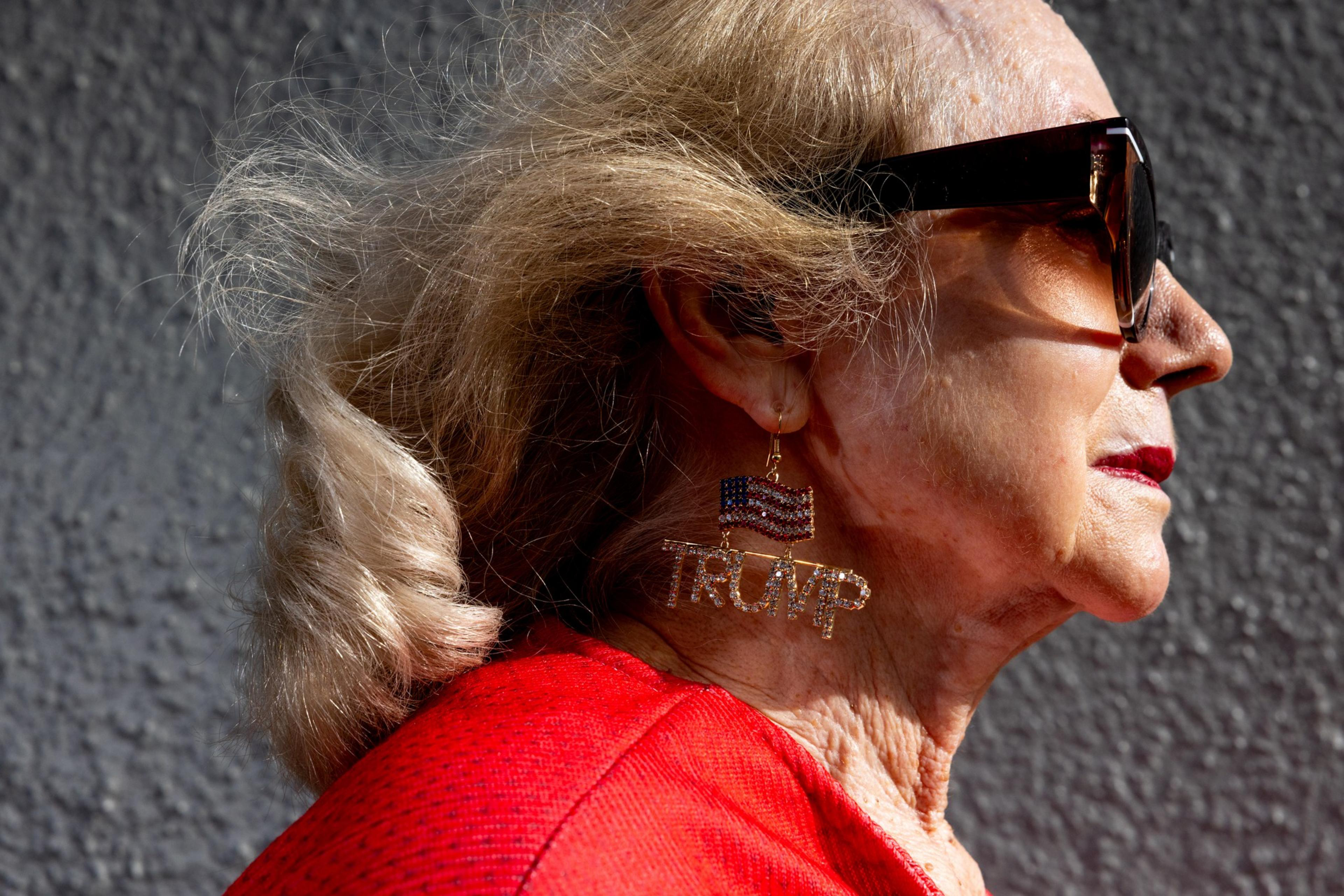 An elderly woman with sunglasses and blonde hair is wearing a red top and an earring featuring an American flag and the word "TRUMP."