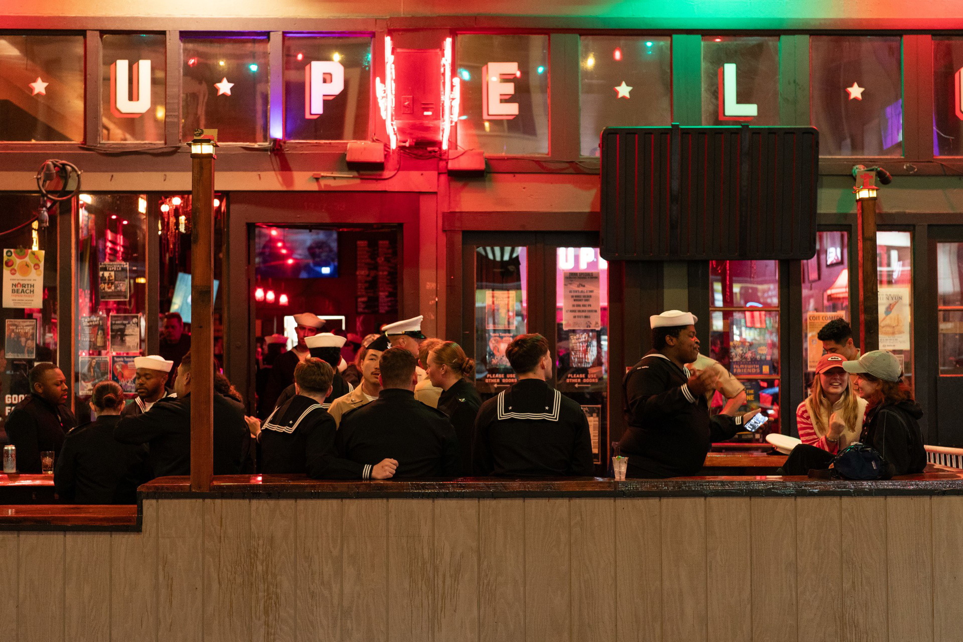 A group of people in navy uniforms converse outside a lively bar with neon signs, surrounded by warm lighting and a casual atmosphere.