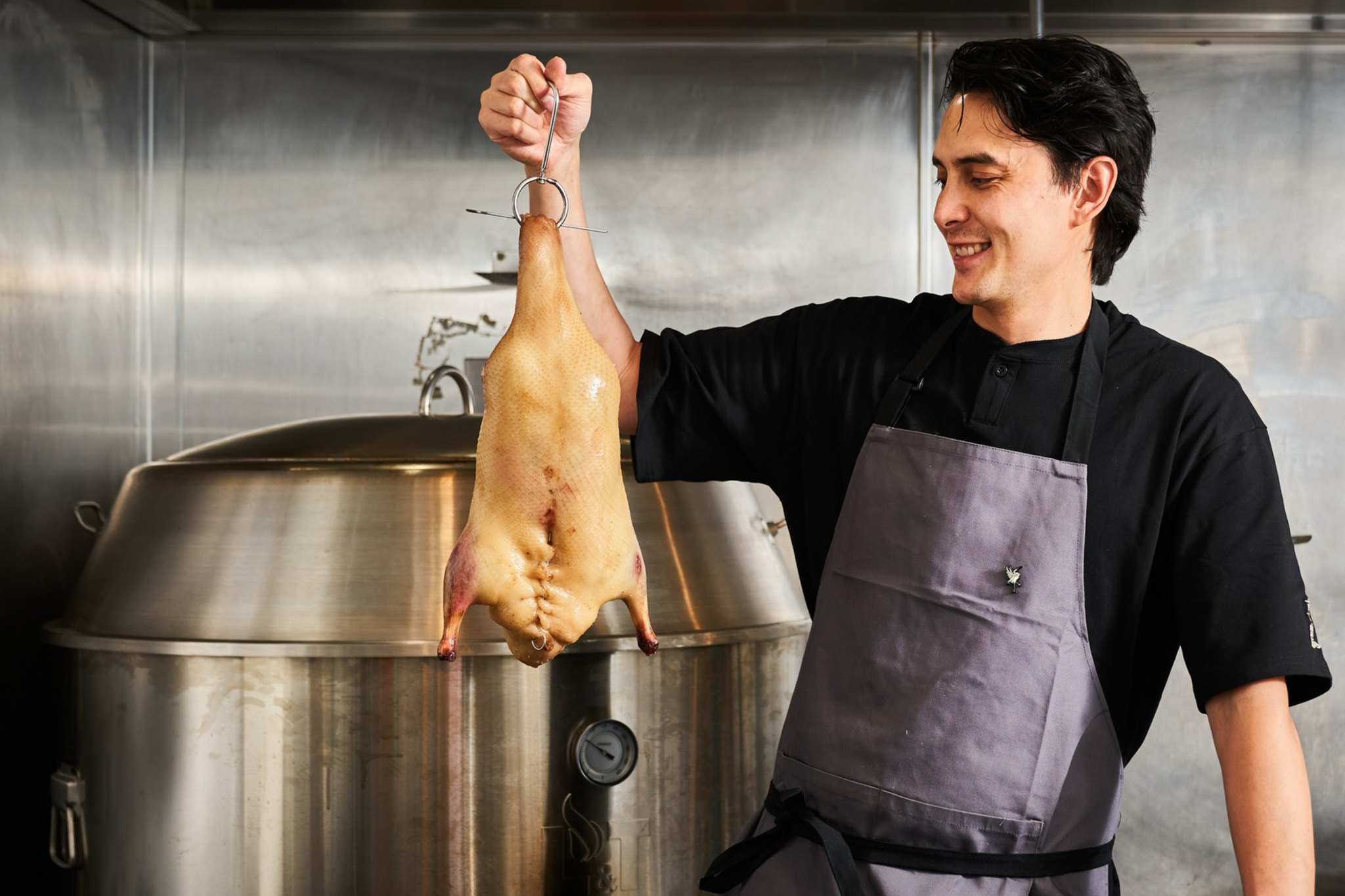 A man in a black shirt and gray apron smiles while holding up a whole plucked duck by a metal hook in a stainless steel kitchen.