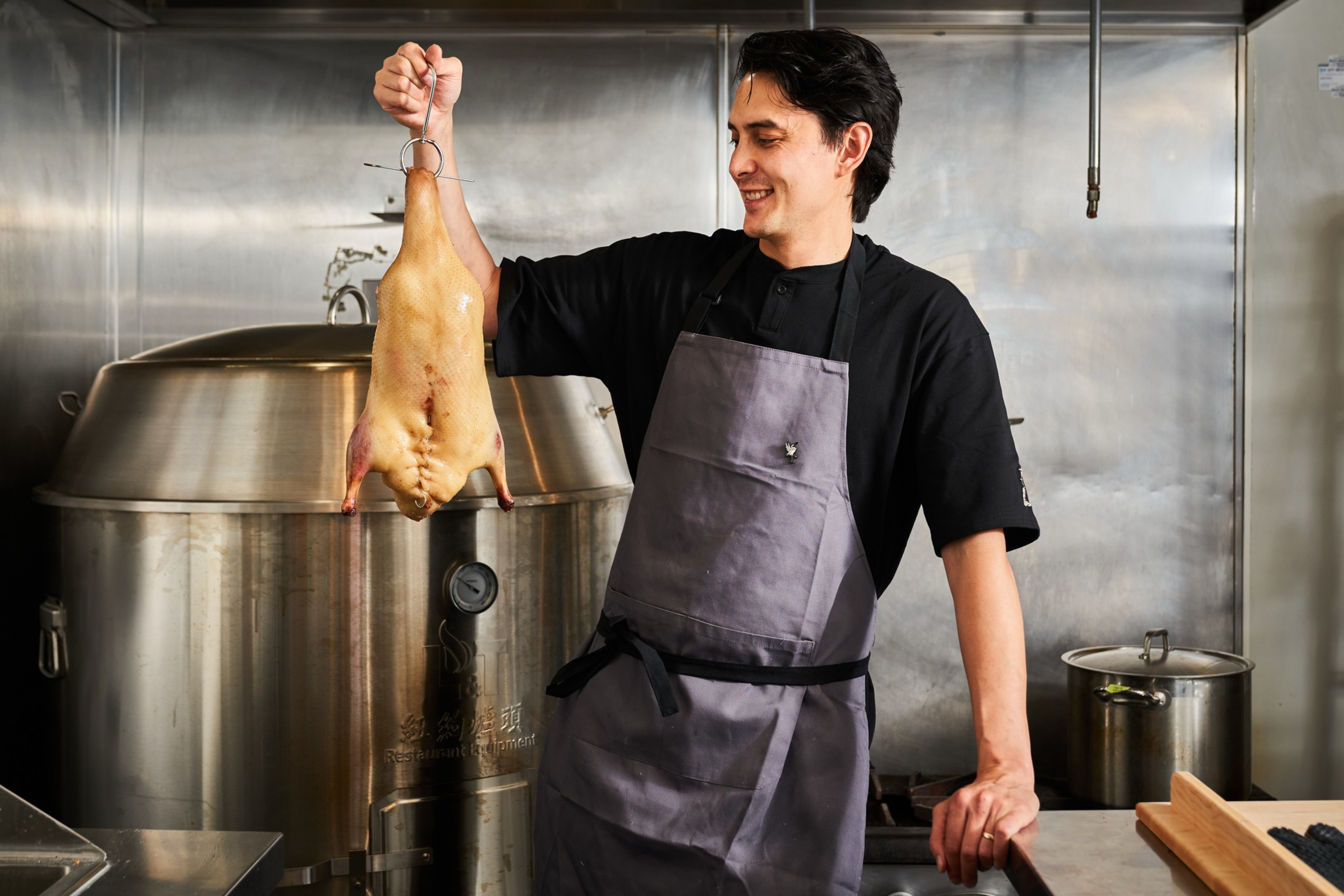 A man in a black shirt and gray apron smiles while holding up a whole plucked duck by a metal hook in a stainless steel kitchen.