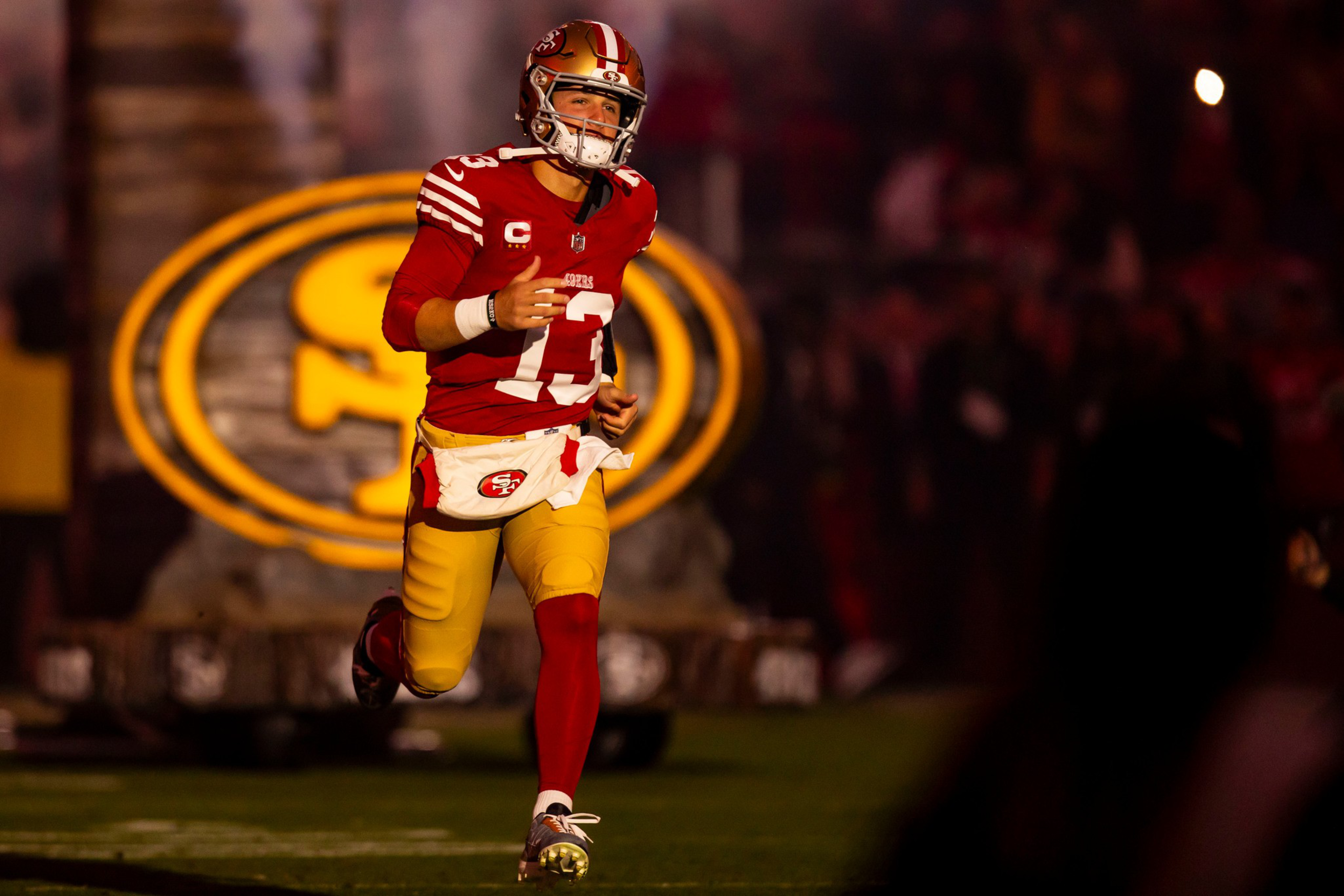 A football player wearing a red San Francisco 49ers jersey and gold pants runs on the field with the team’s logo illuminated behind him.