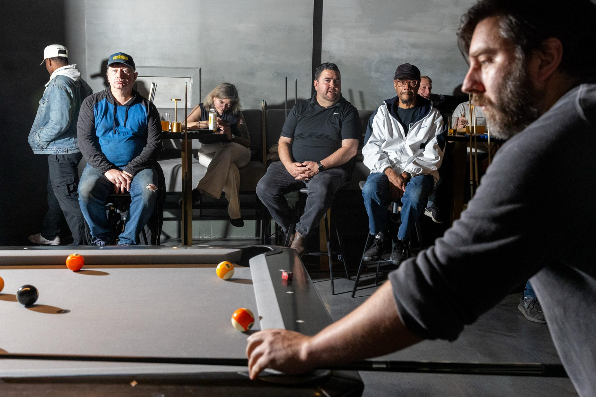 A man focuses on taking a shot in pool while five people sit and watch from behind in a dimly lit room.