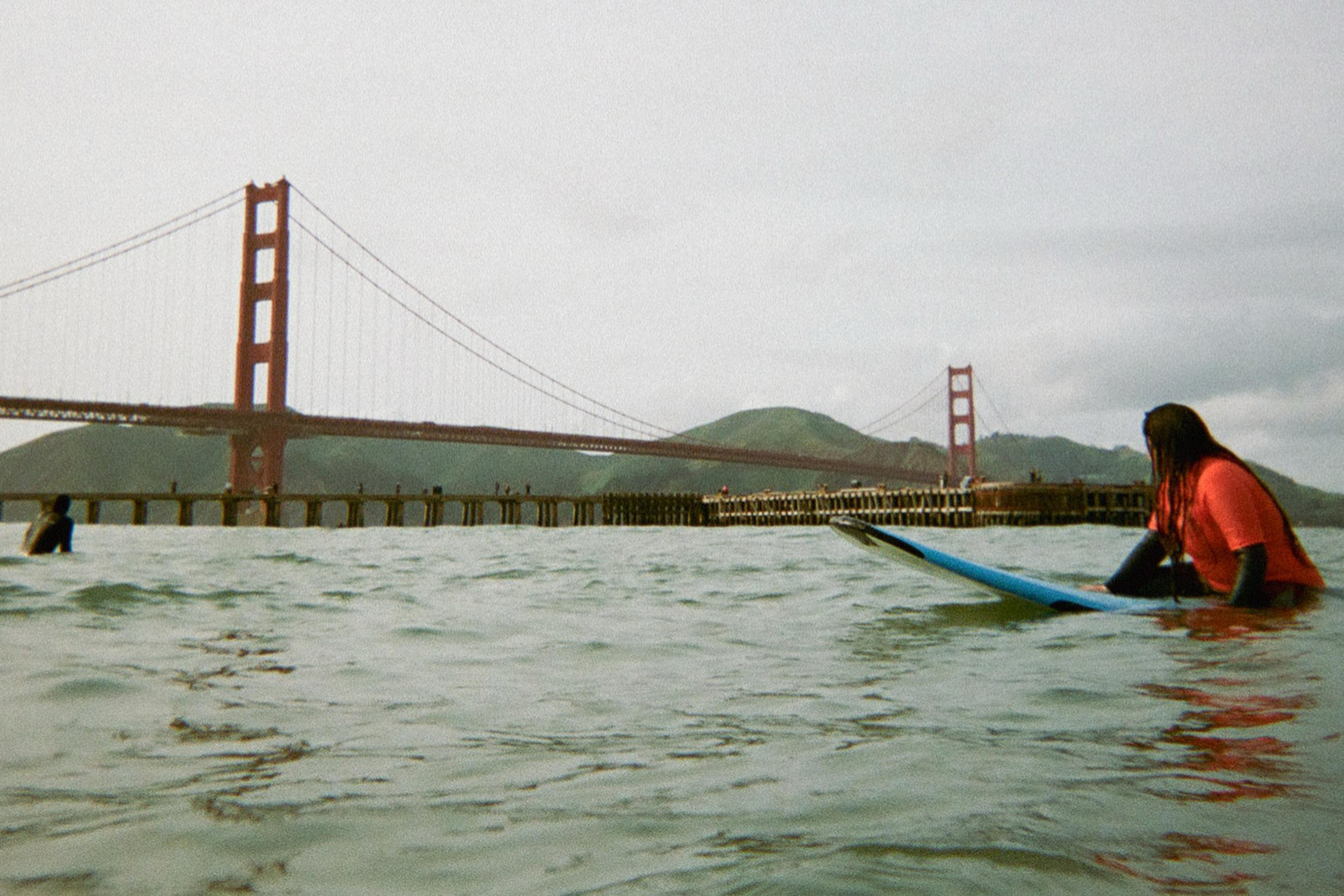 A person on a surfboard sits in the water near the Golden Gate Bridge, with hills in the background and a pier extending out into the water.