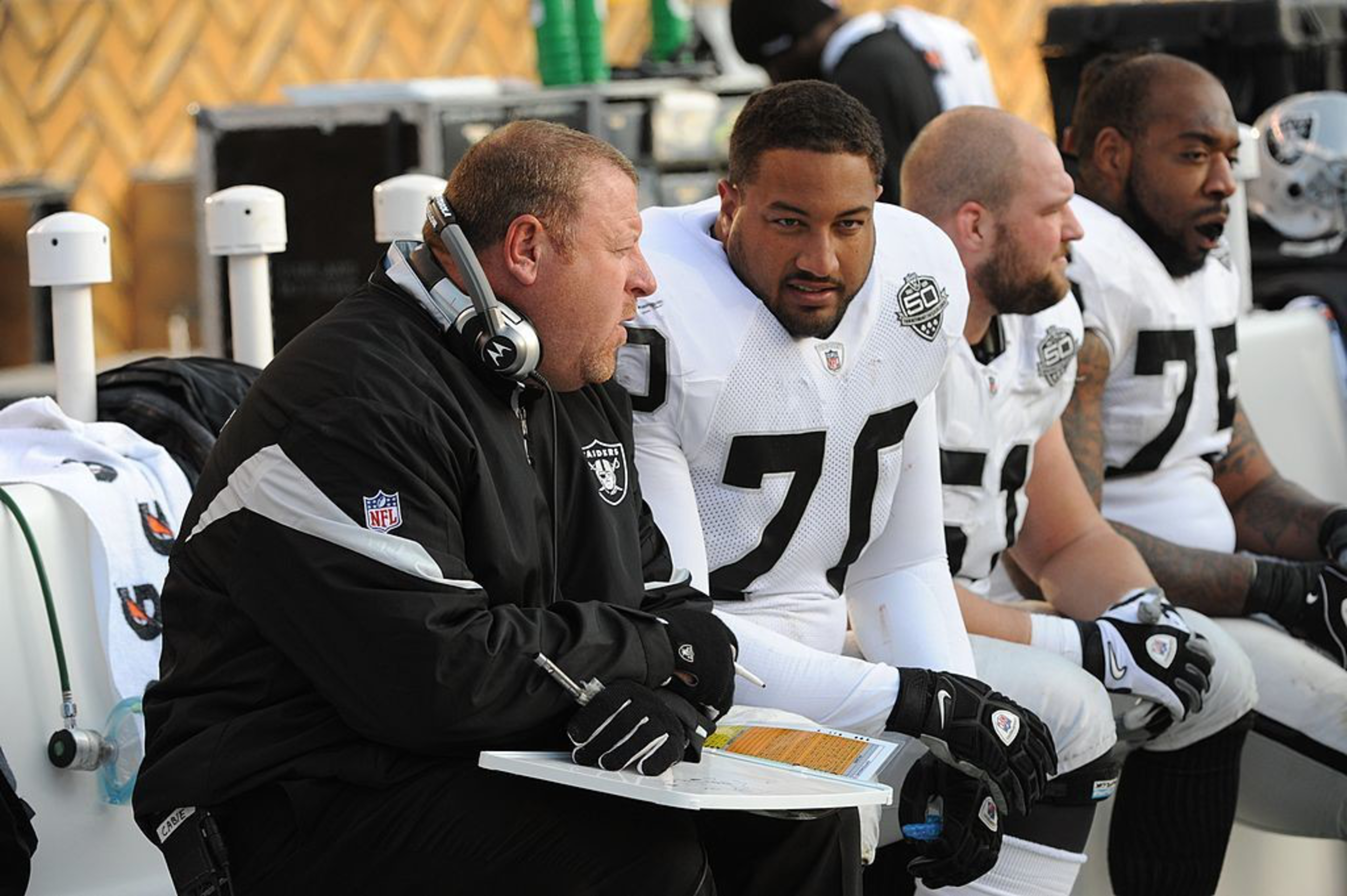 A coach discusses strategy with three football players on the sideline during a game. They're focused and wearing team gear.