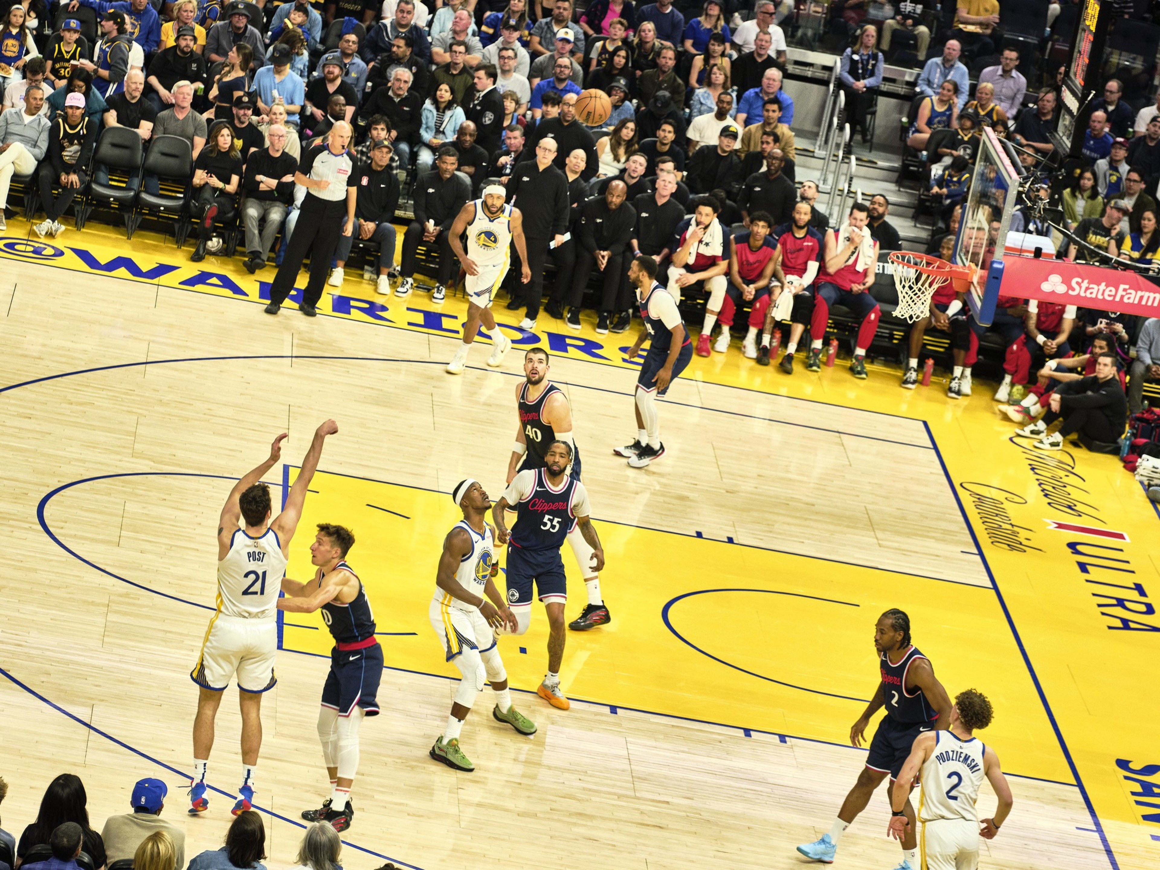 A basketball player in a white jersey takes a jump shot while others watch. The court is vibrant with a yellow and blue design, and the stands are full of spectators.