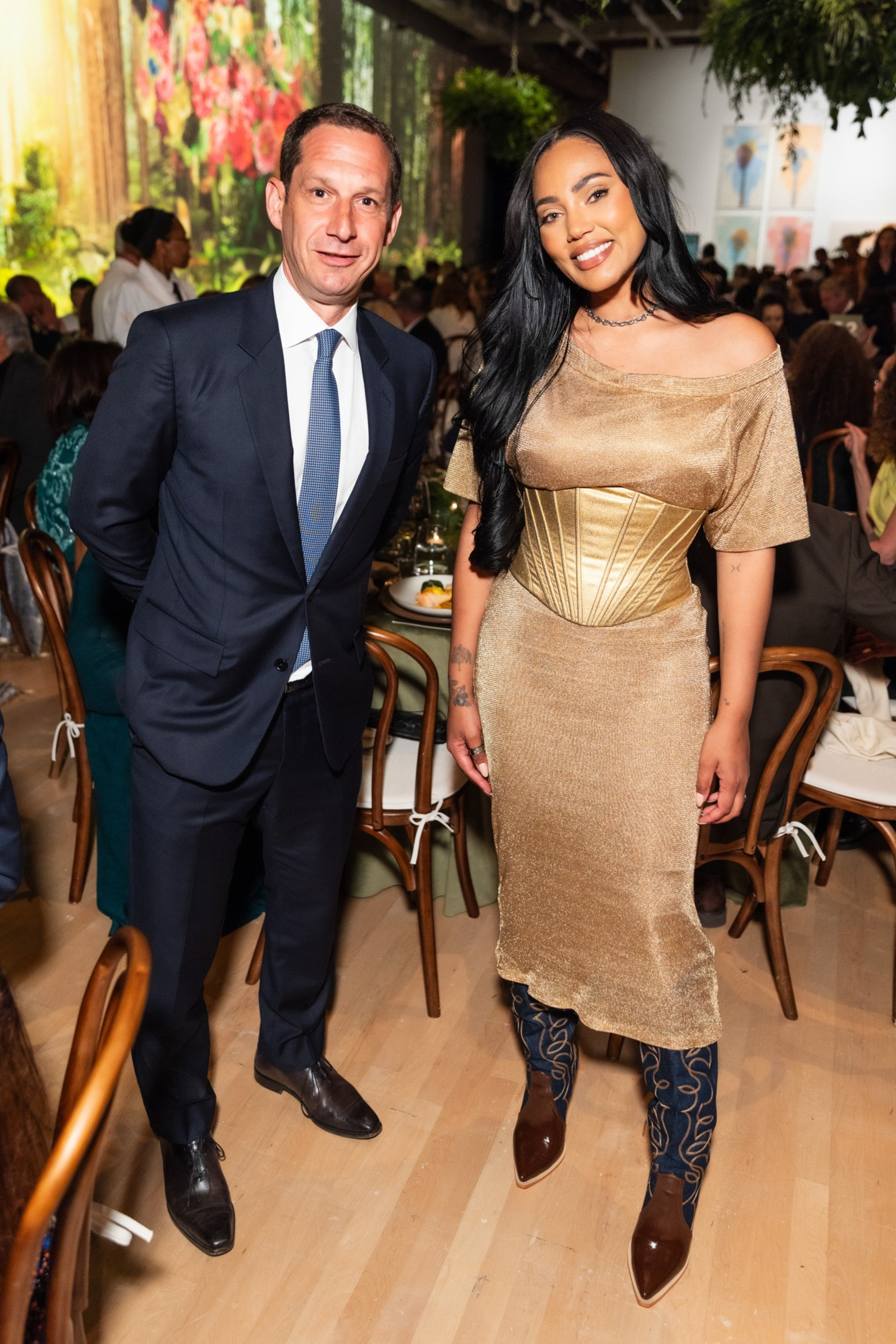 A man in a suit and a woman in a gold dress stand smiling in a crowded room with wooden chairs and a floral backdrop.