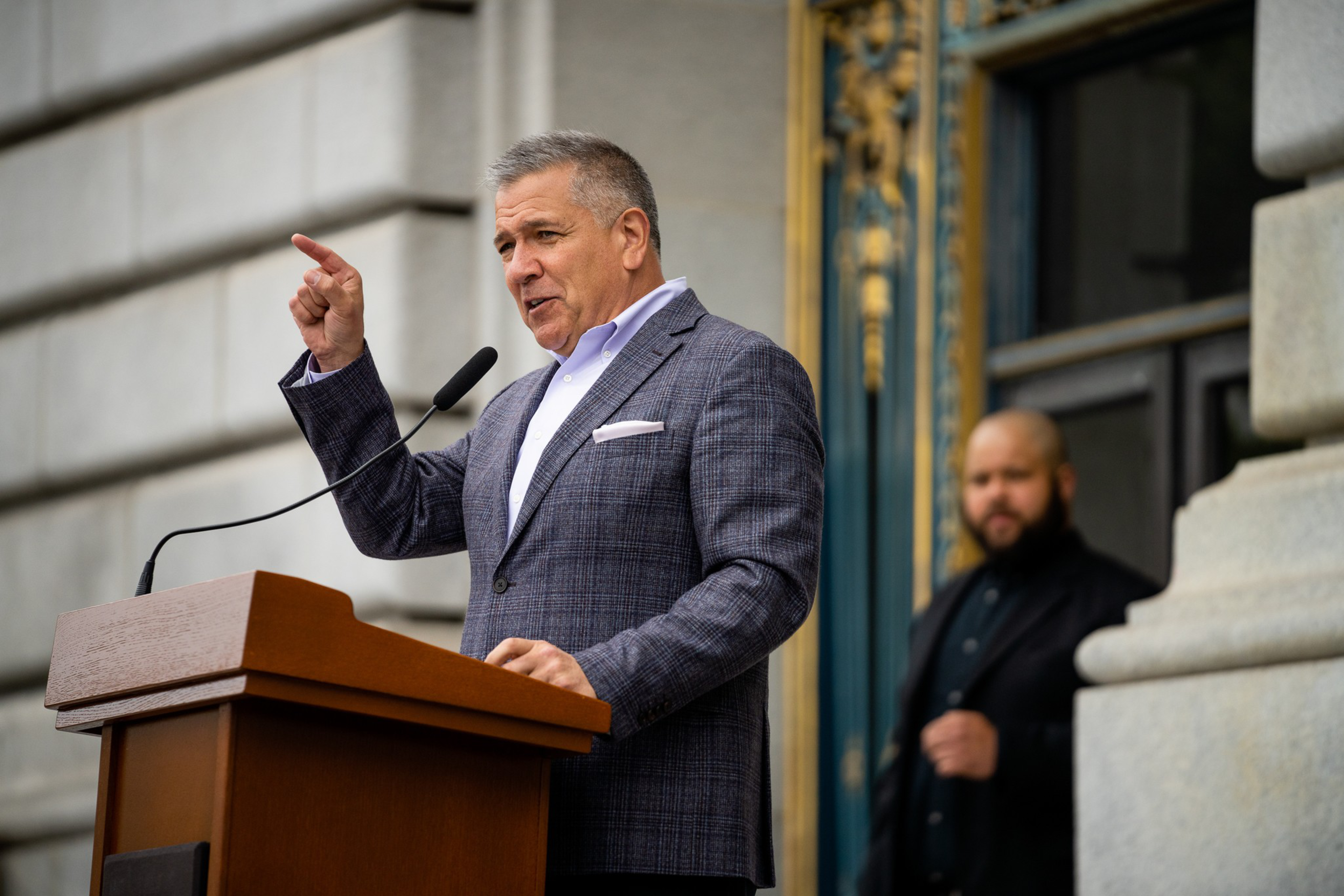 A man in a suit speaks passionately at a podium, pointing his finger. Another person stands behind him near a building with ornate architectural details.