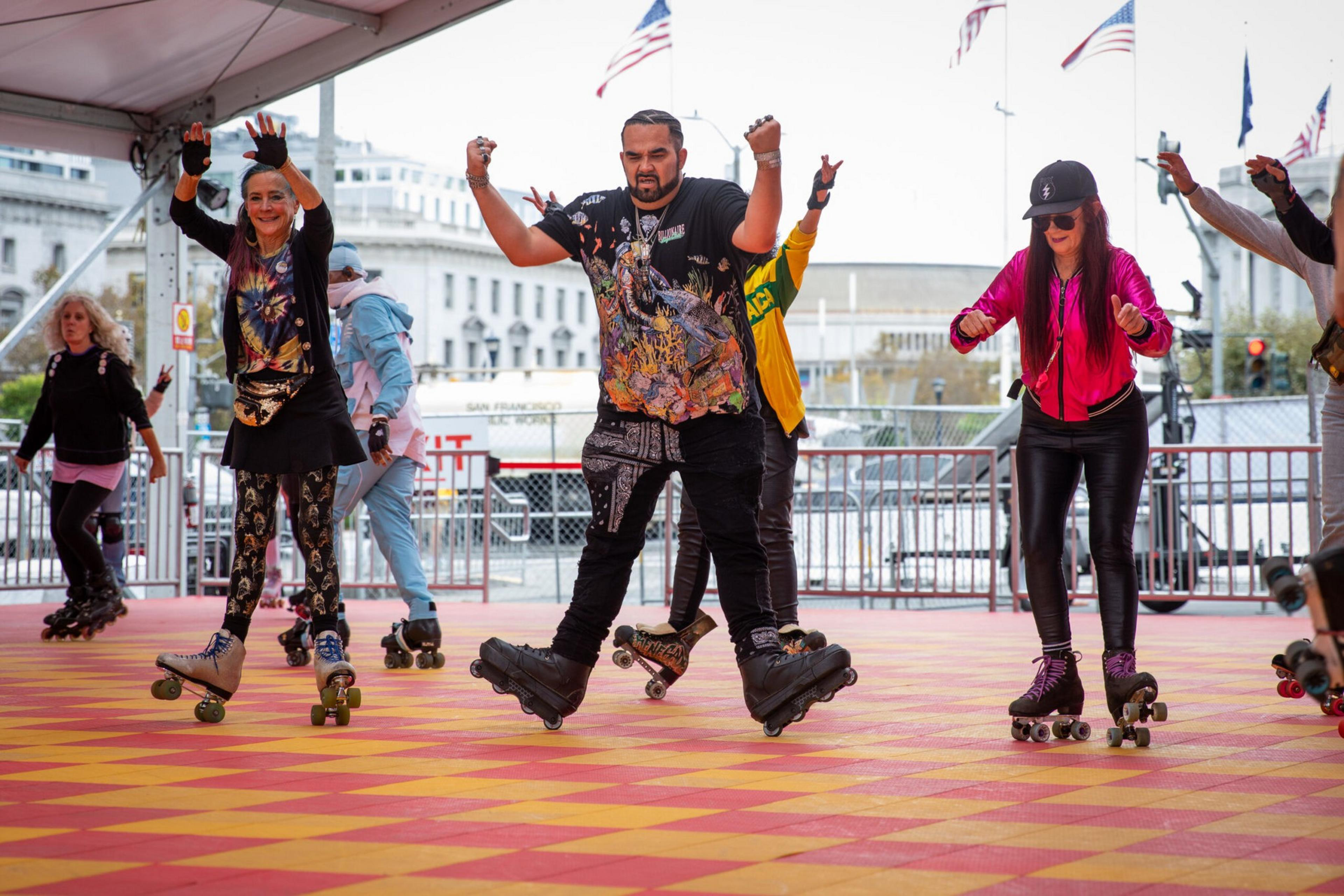 A group of diverse people roller skating energetically on a red and yellow checkered floor under a tent, with American flags and buildings in the background.
