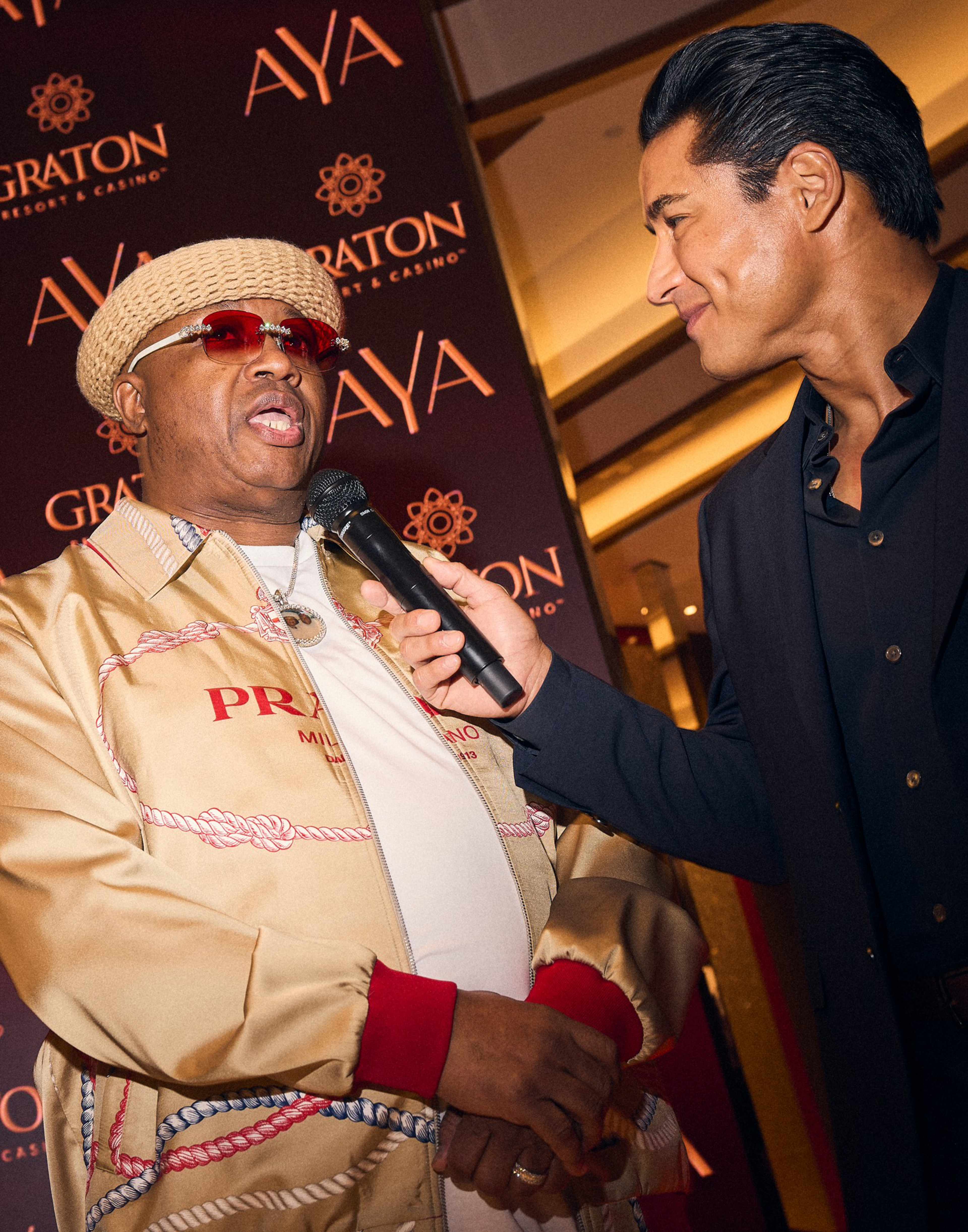 A man in a beige Prada jacket and beige knit hat is speaking into a microphone held by another man in a dark suit smiling at him.