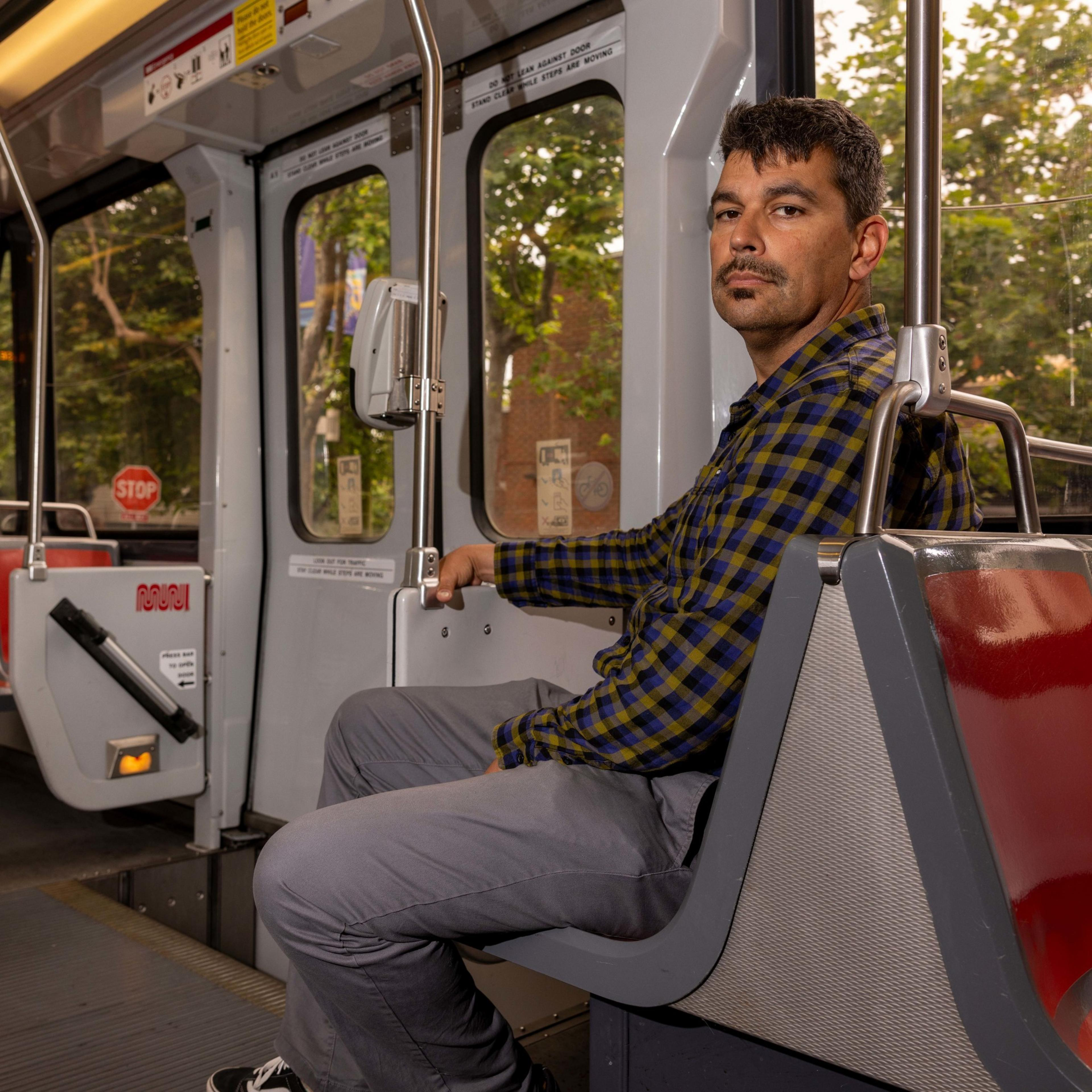 A man with short hair and a mustache, wearing a plaid shirt and gray pants, sits on a public transit seat.