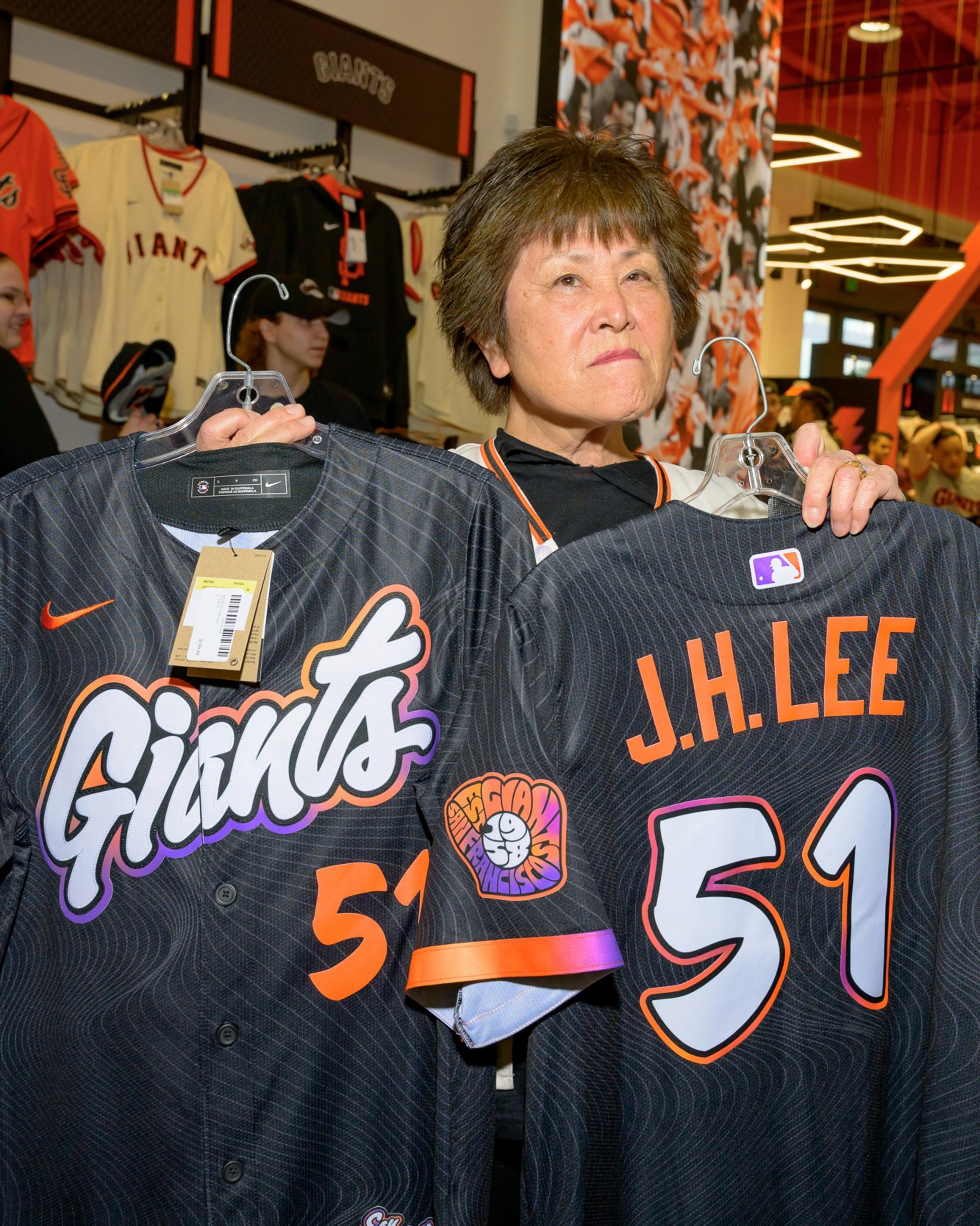 An individual holds two black jerseys with "Giants" and number 51 printed. One jersey shows "J.H. Lee" on the back. Other jerseys hang in the background.
