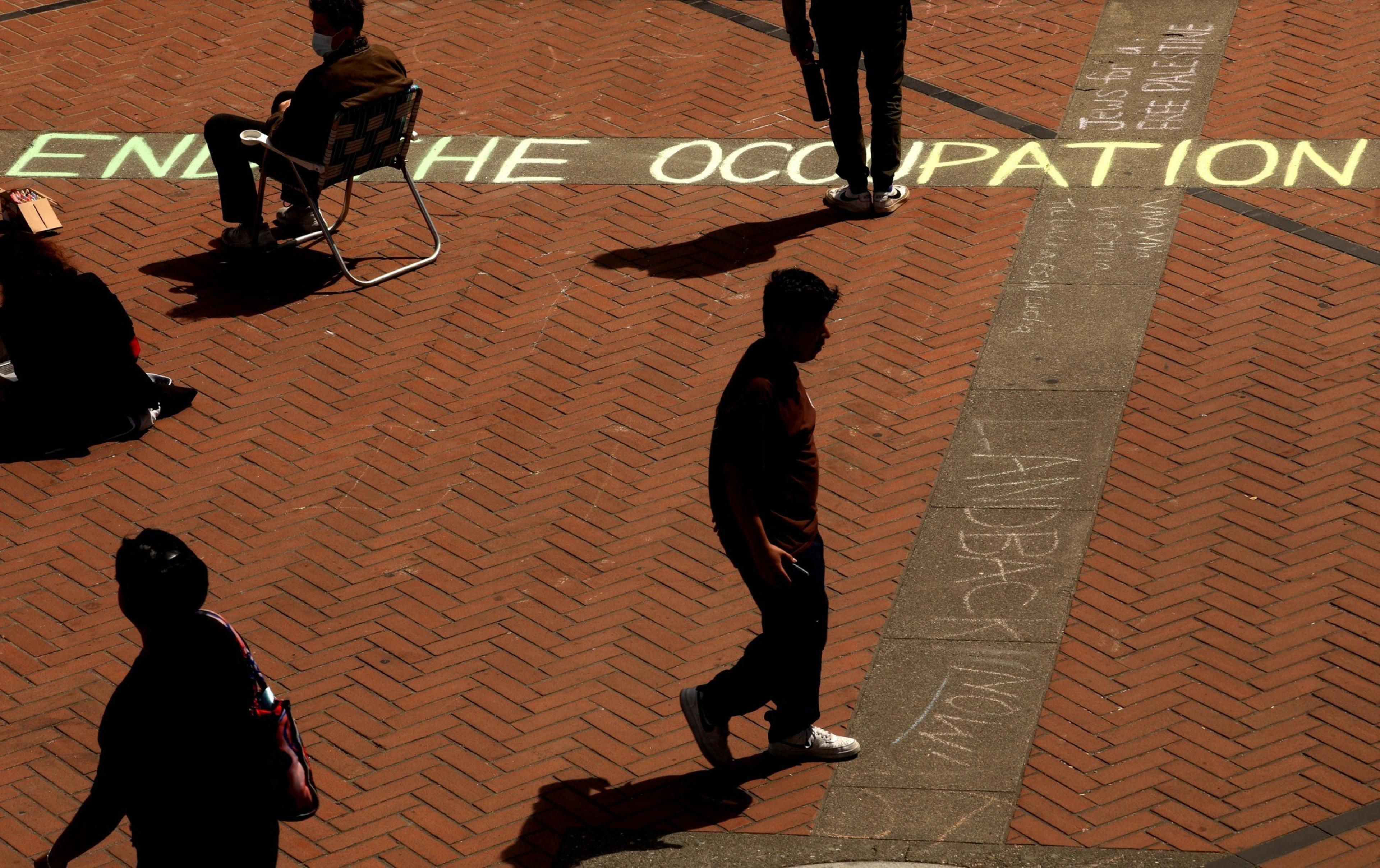 People walk over brick pavement with the phrase &quot;END THE OCCUPATION&quot; chalked in large letters. Shadows suggest bright sunlight.