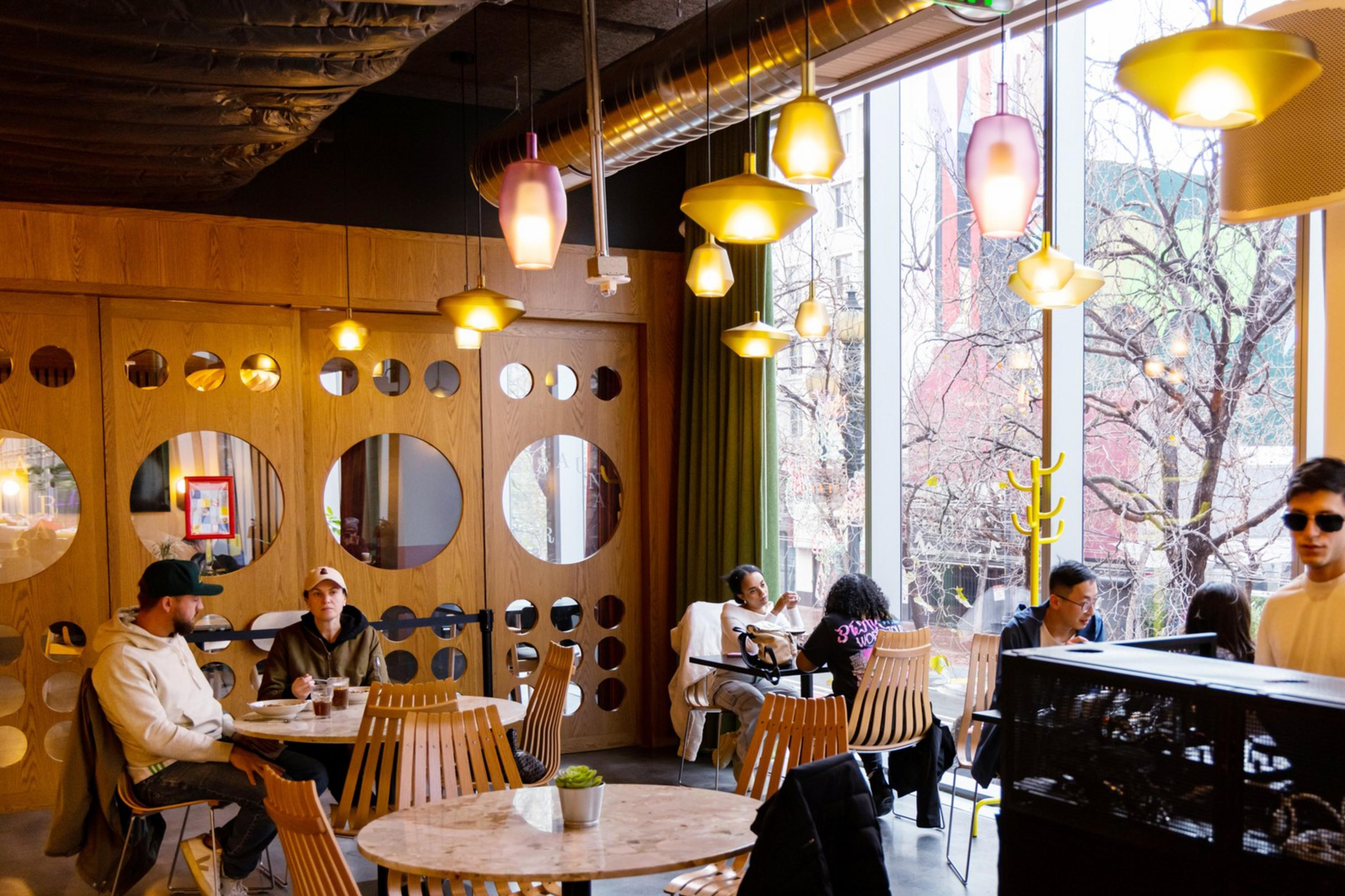 A cozy cafe with wooden decor features people sitting at tables, chatting and using laptops. Two individuals are at a DJ booth near large windows.