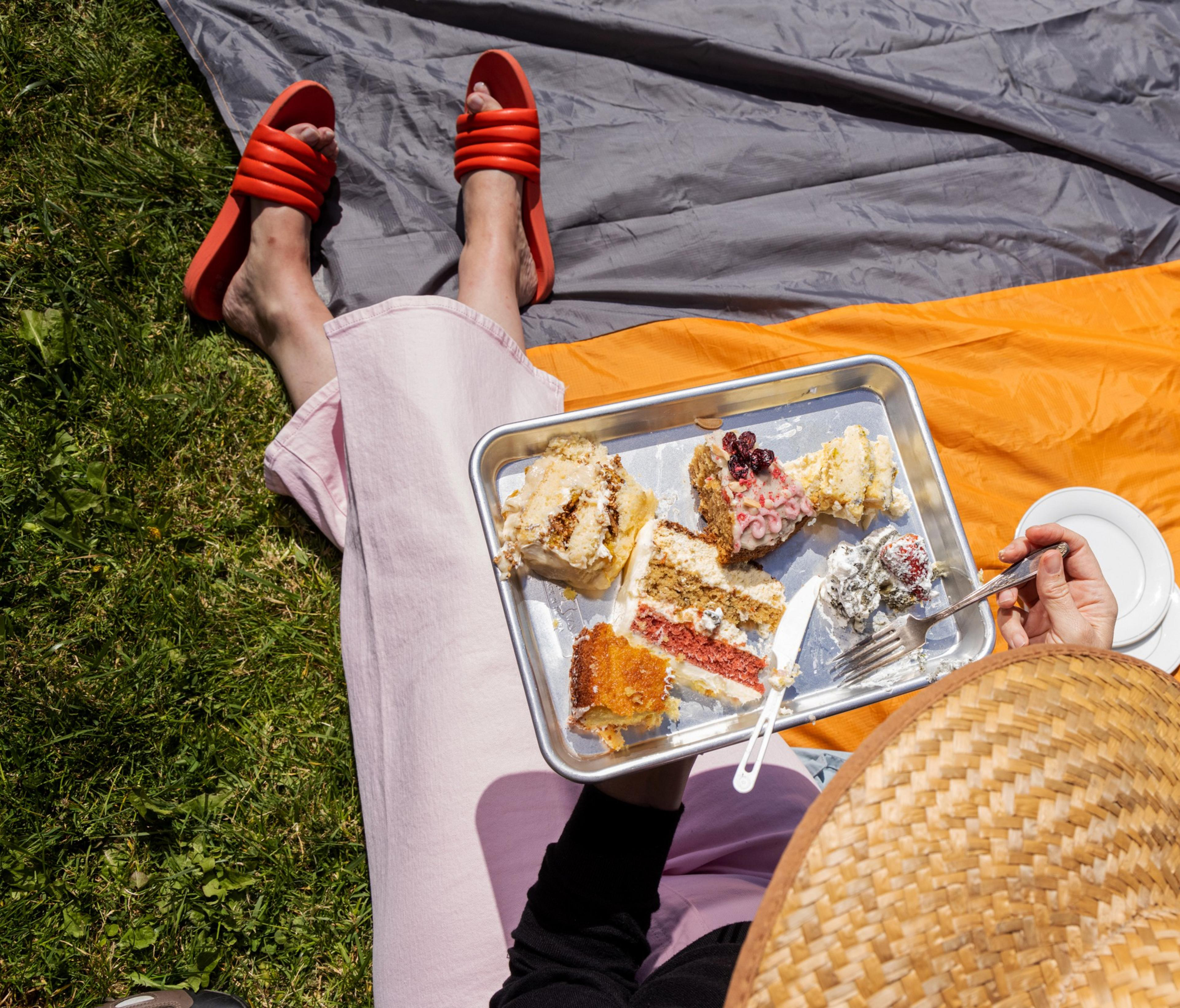 A person with pink pants and red sandals is sitting on grass, holding a tray of assorted cake slices. They're also holding a straw hat and a fork.