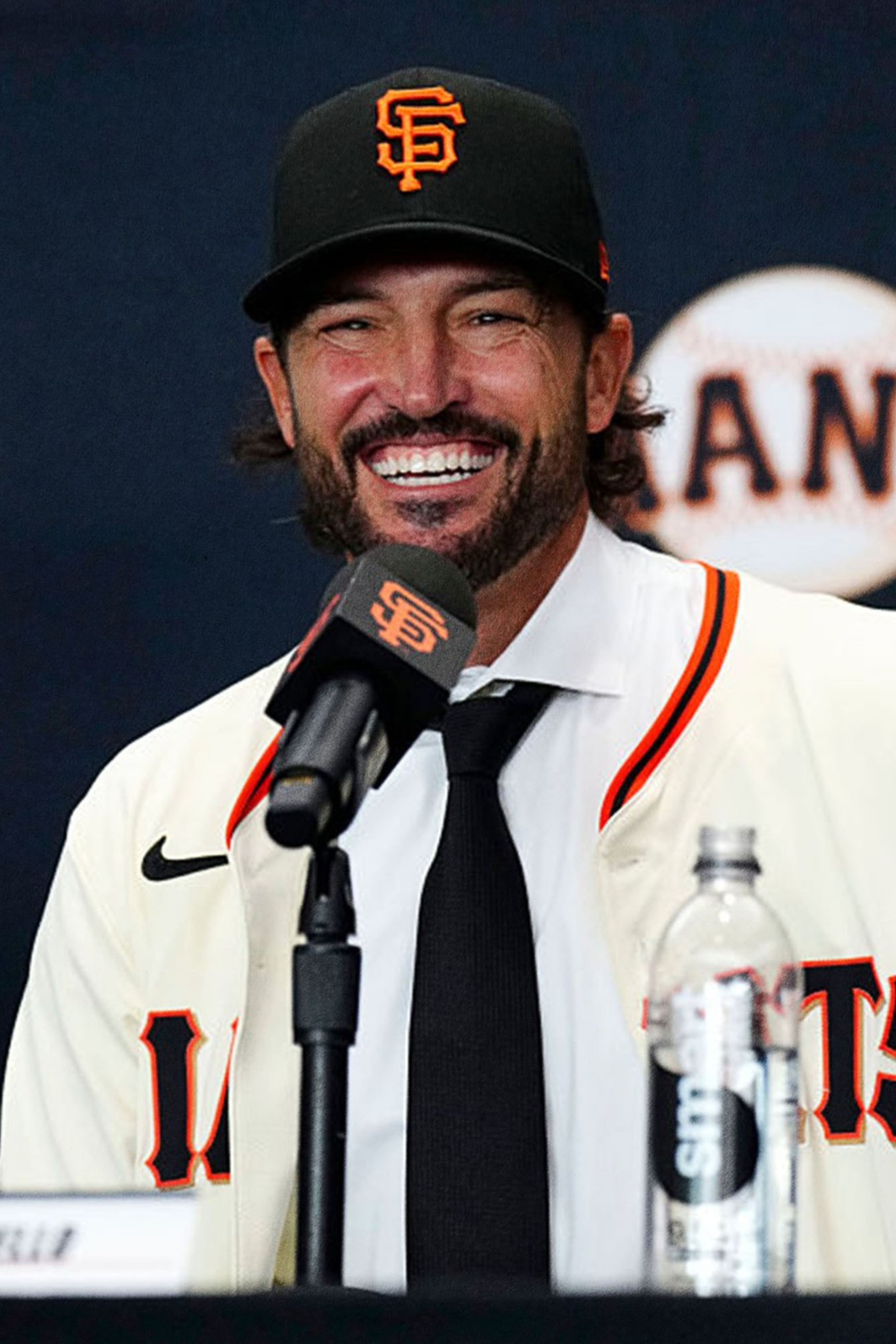 A smiling man wearing a San Francisco Giants baseball cap and jersey sits at a press conference microphone, with baseball-themed images in red and black on the side.