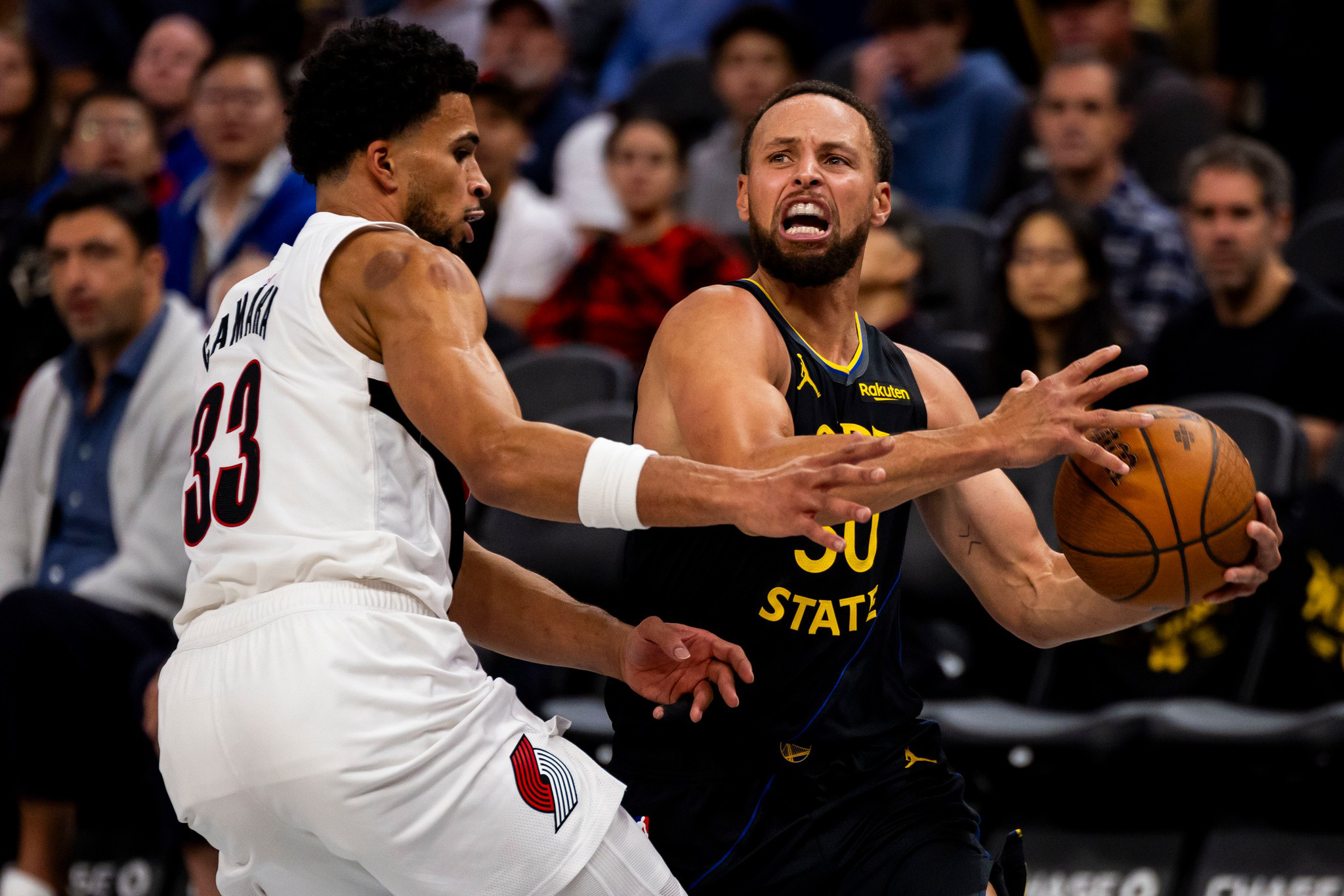 Stephen Curry, wearing a Golden State Warriors jersey, tightly grips a basketball while being closely defended by a player in a white Portland Trail Blazers uniform.