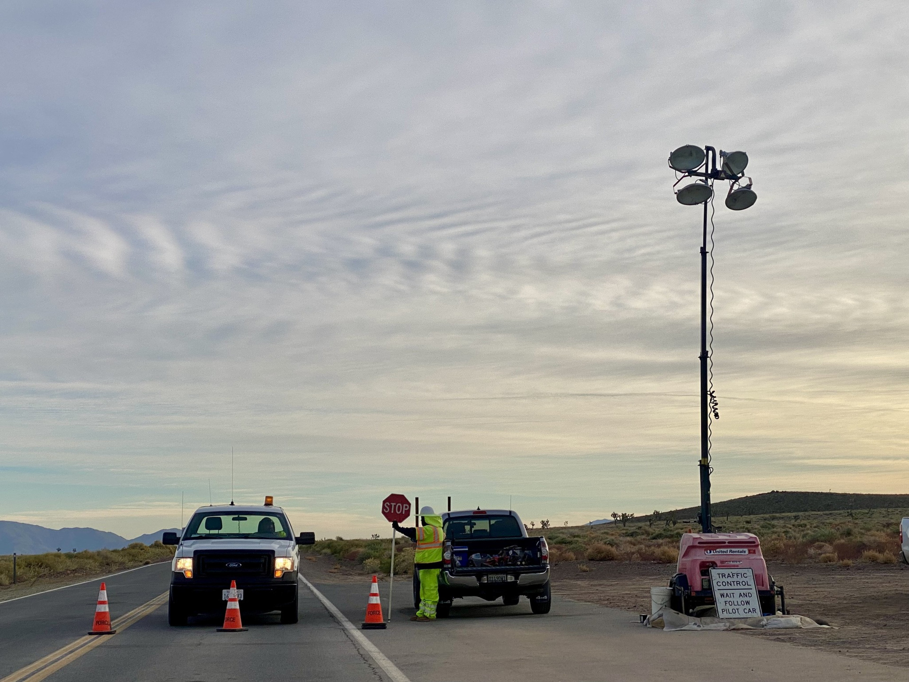 Two pickup trucks behind orange cones block a roadway, where a person in neon yellow workwear holds up a stop sign. 