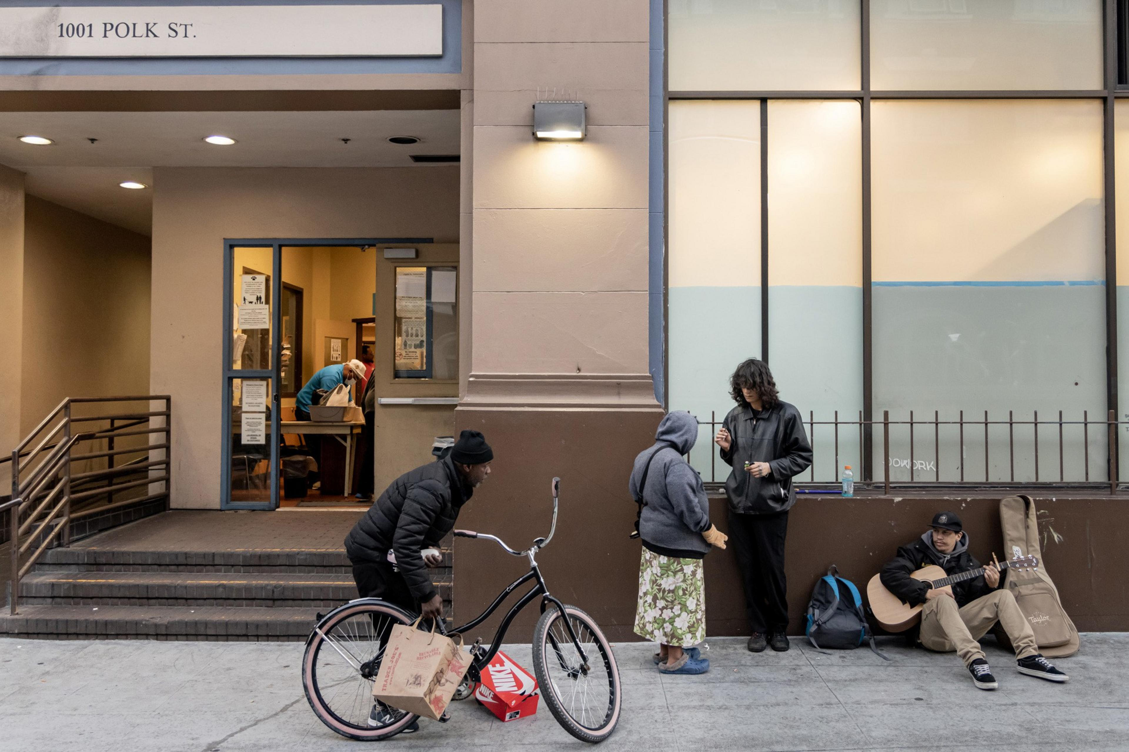 People are gathered outside a building at 1001 Polk St. One rides a bike holding a bag, two are talking, and another sits playing guitar. A person is seen inside.