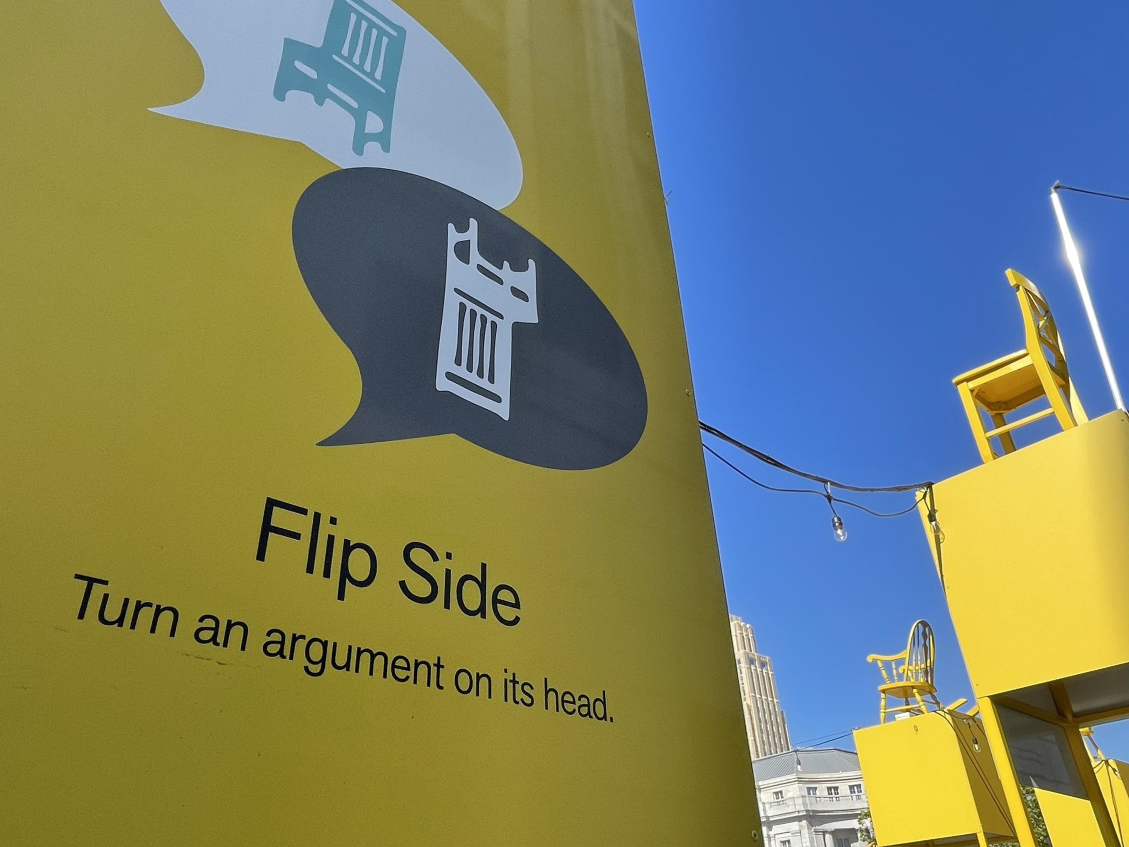 A yellow sign with text &quot;Flip Side Turn an argument on its head,&quot; two inverted speech bubbles with chair icons, under a blue sky.