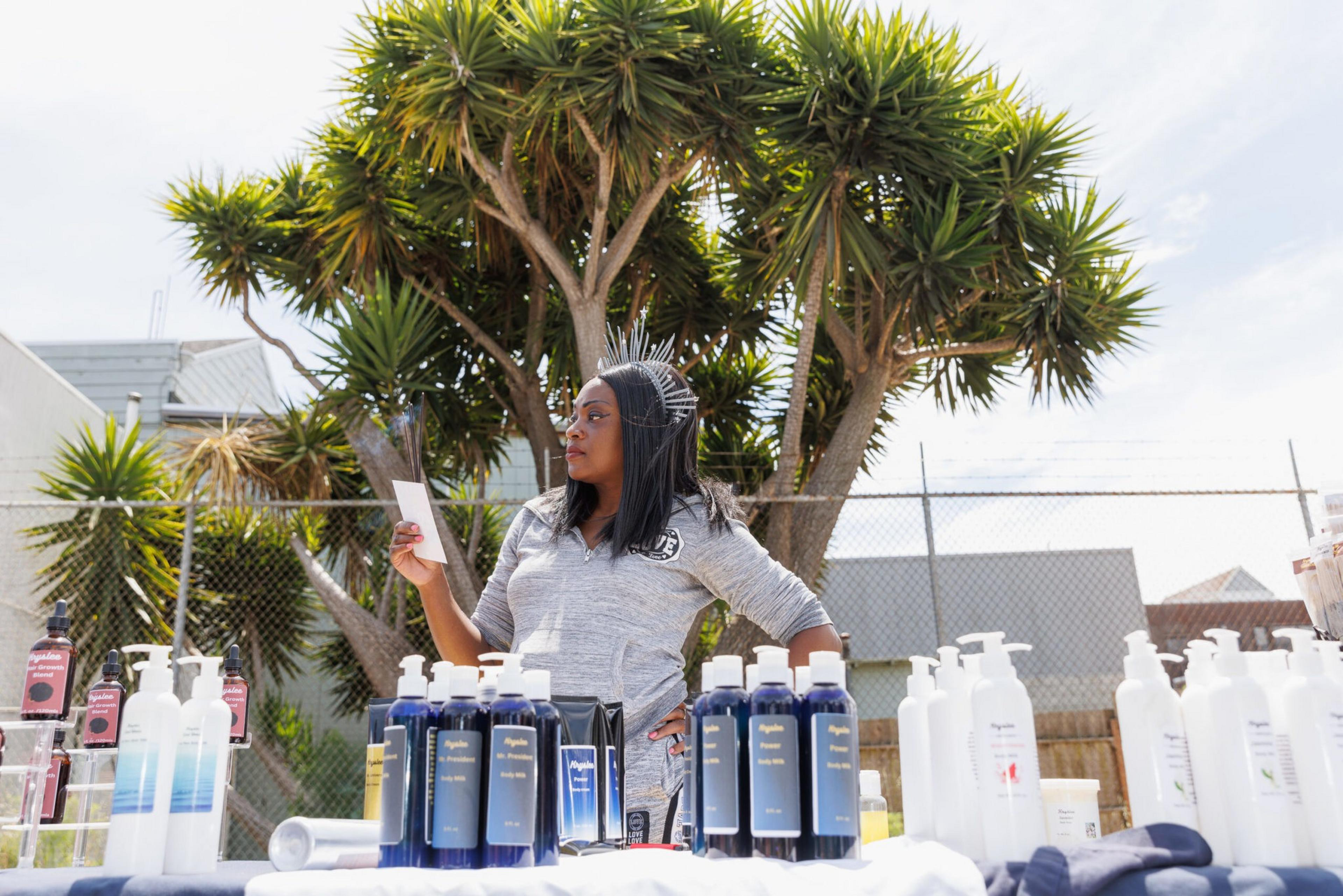 A woman stands behind a table with hair care products, palm trees in the background.