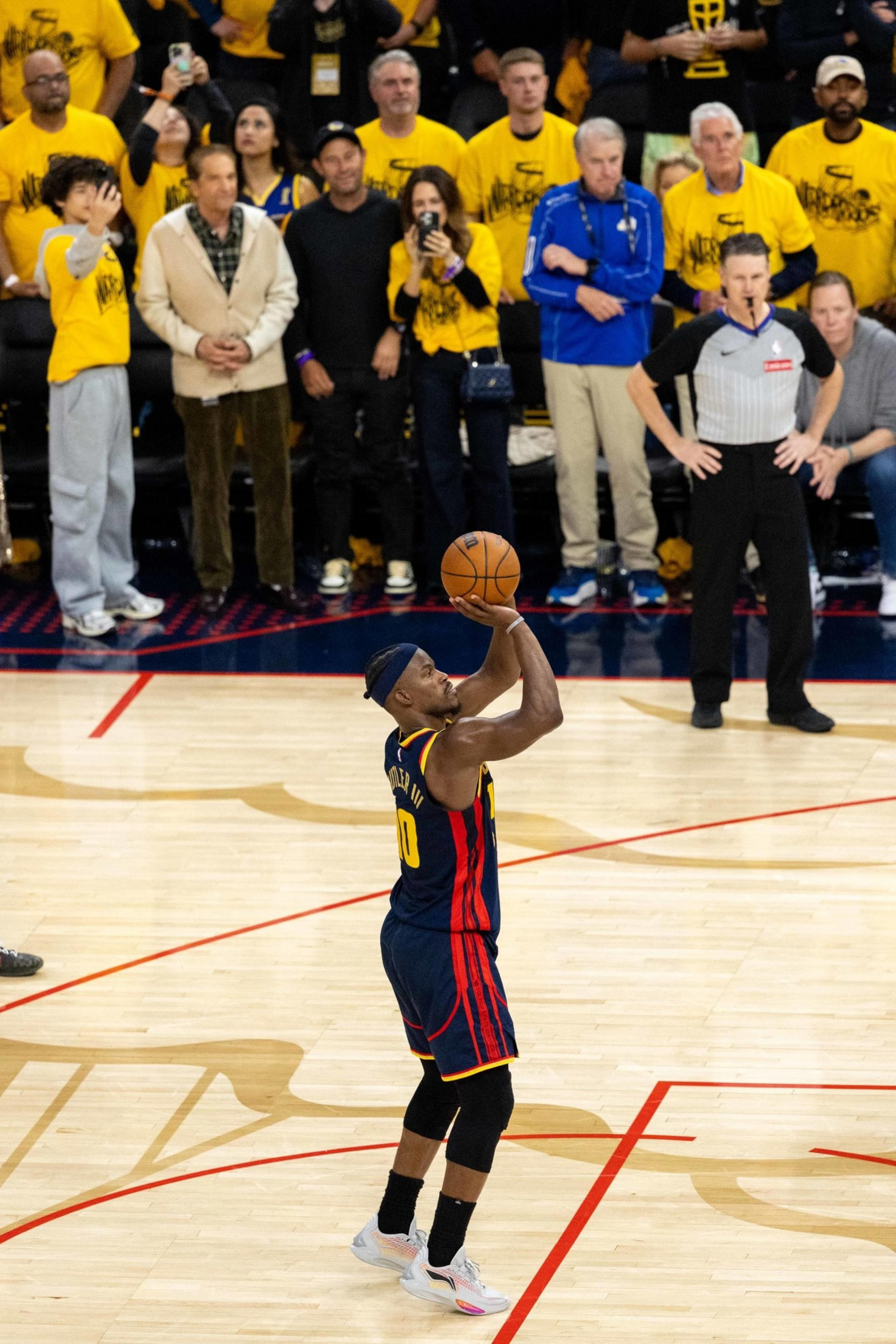 A basketball player in a navy jersey takes a free throw shot on a court, surrounded by a crowd in yellow shirts. An official observes closely.