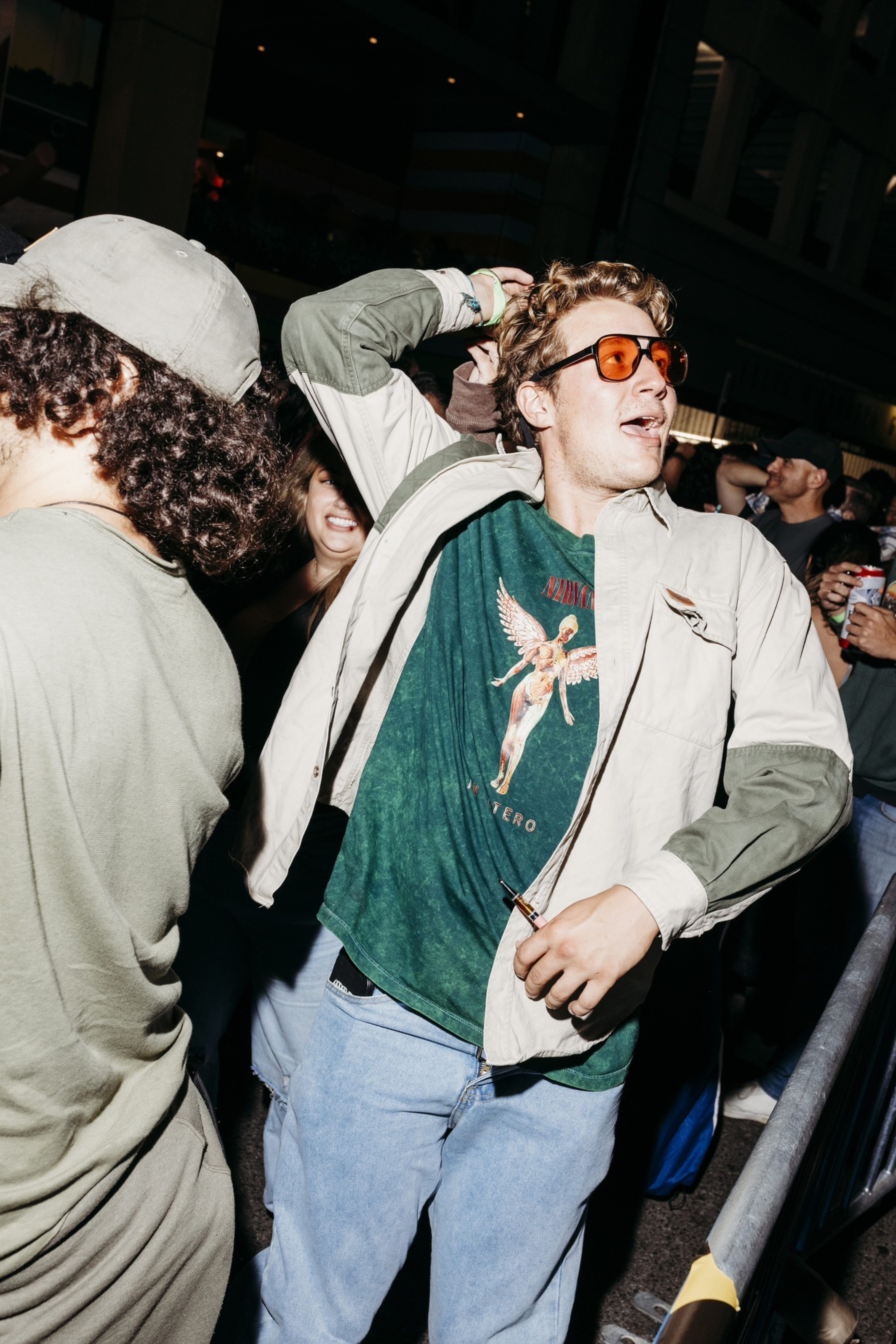 A young man wearing orange sunglasses and a graphic tee dances energetically at a crowded nighttime event with other casually dressed people.