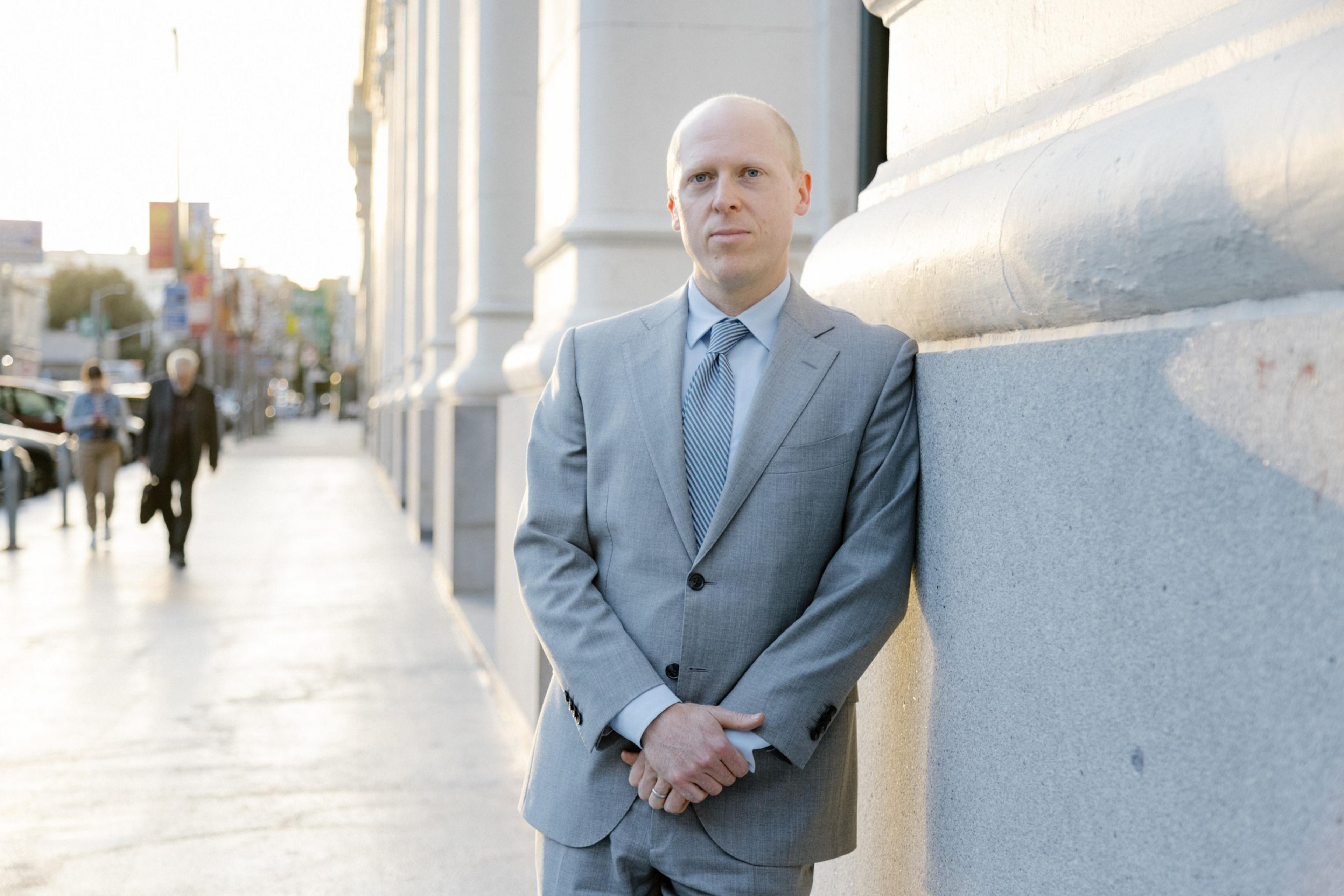 A man in a gray suit stands by a street, leaning against a building, with pedestrians in the soft-focus background.